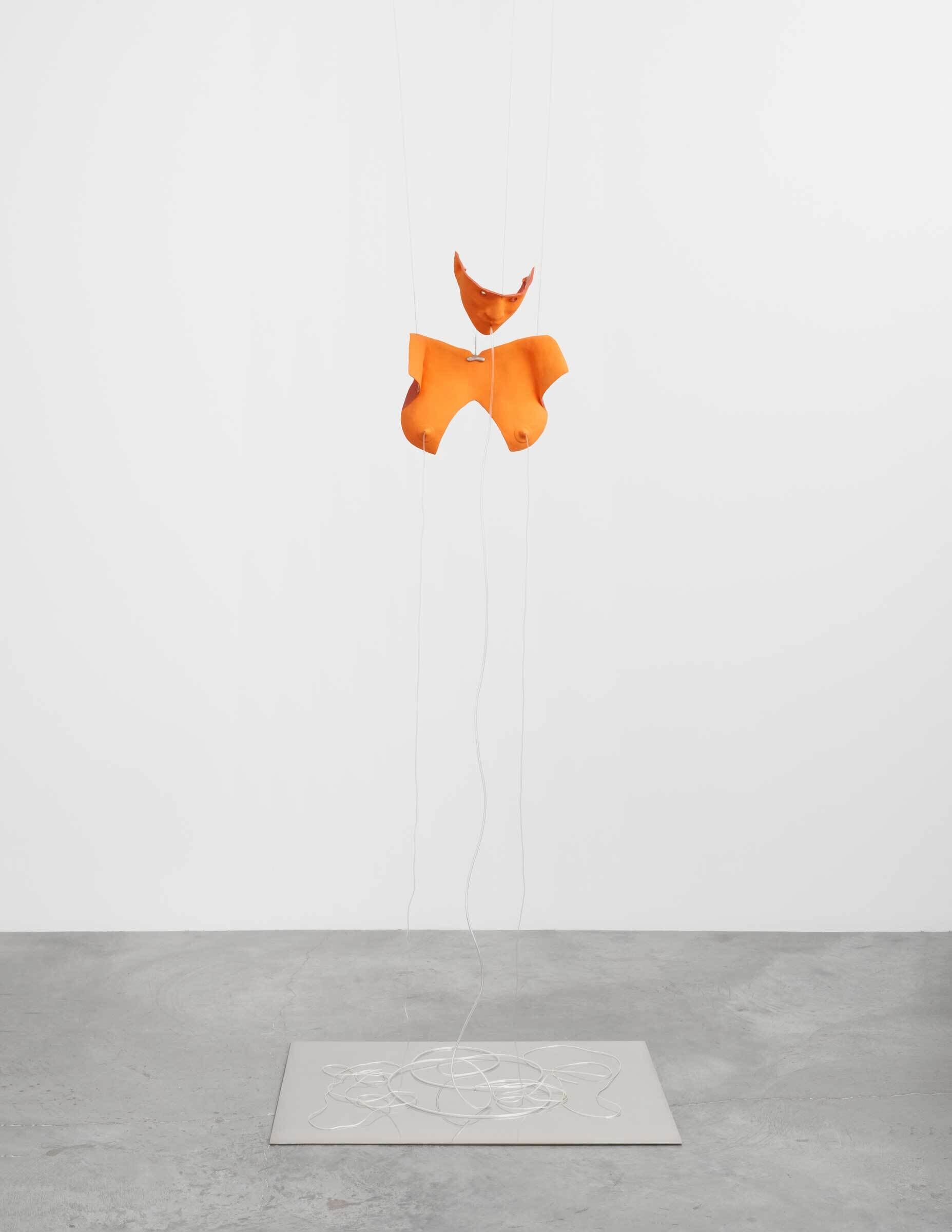 An orange abstract sculpture suspended by strings above a white outline on the floor in a minimalist setting.