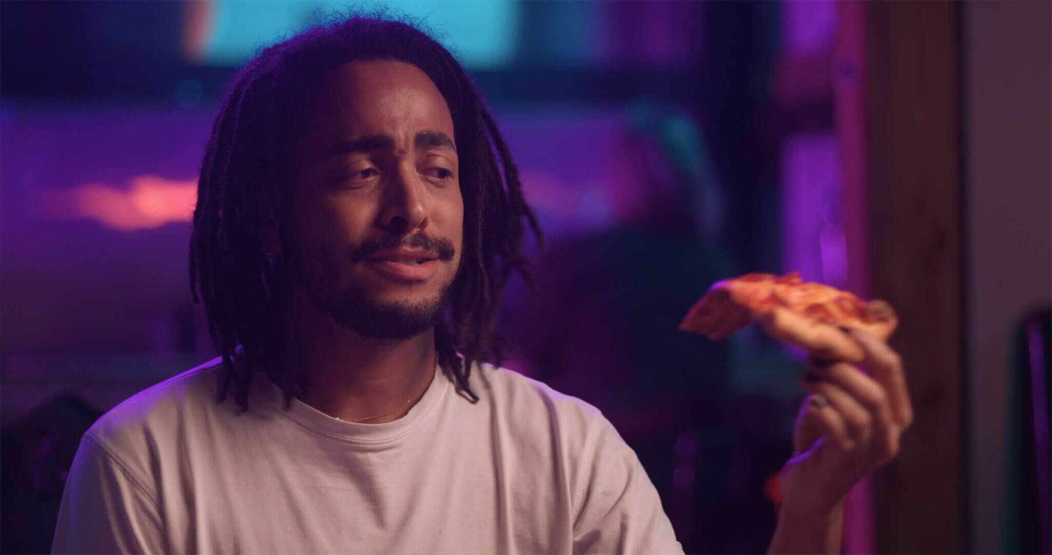 Man with dreadlocks holding a slice of pizza, looking content in a room with purple lighting.