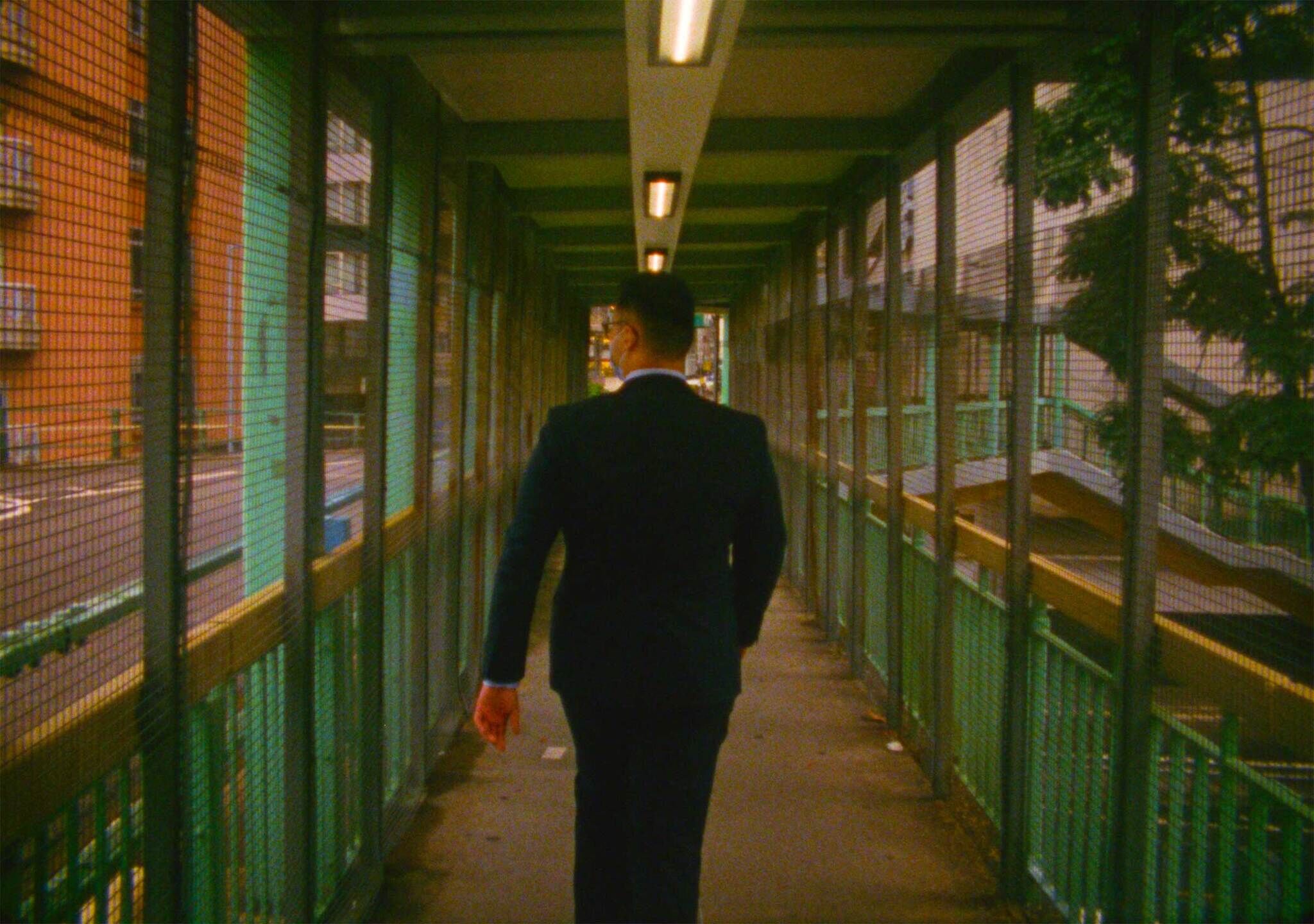 Man in suit walking down a covered pedestrian bridge with mesh walls and overhead lighting.