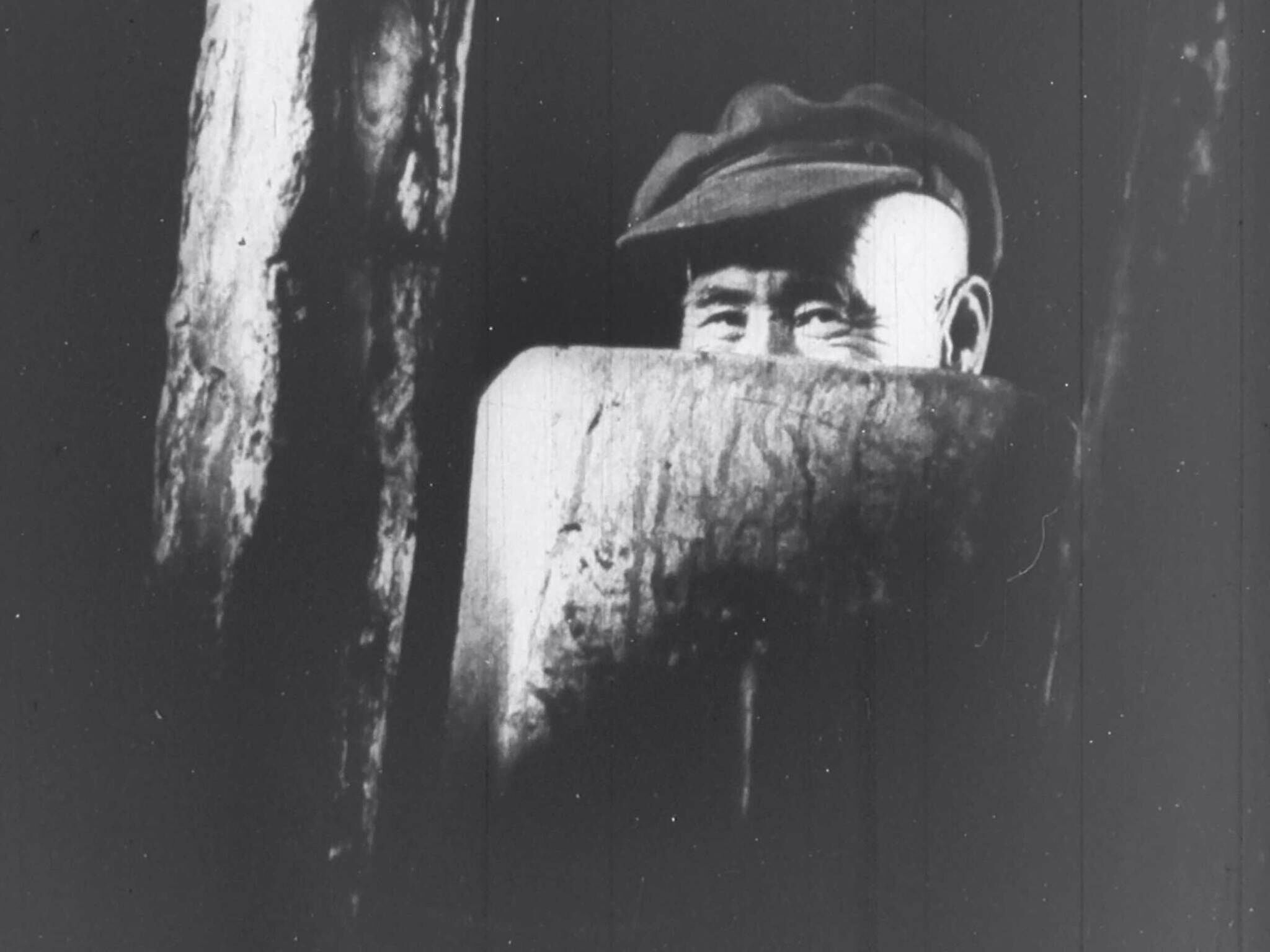 Man in a flat cap peeking through a hole in a wooden fence.