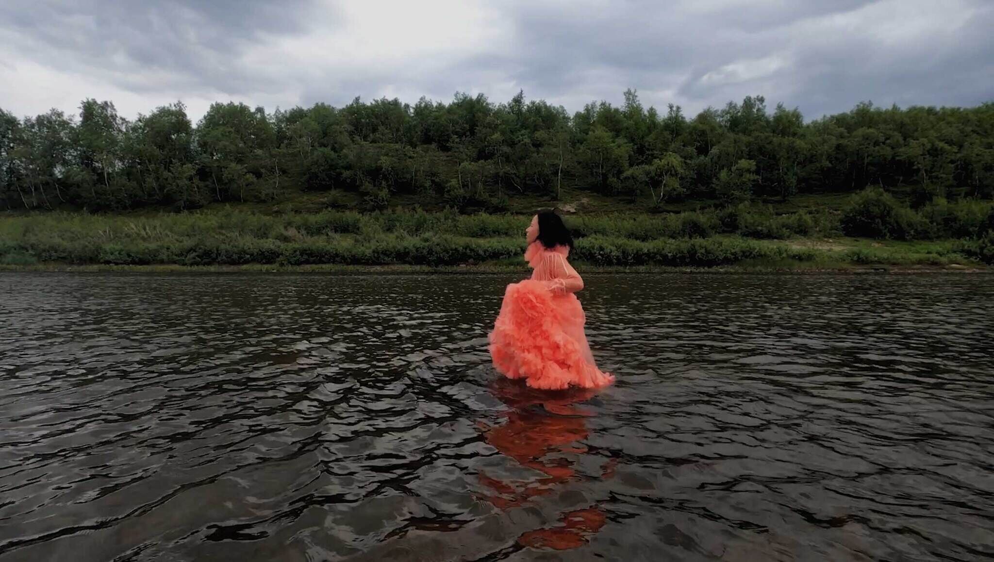 Person in a voluminous orange dress standing in a river with a backdrop of greenery and overcast sky.