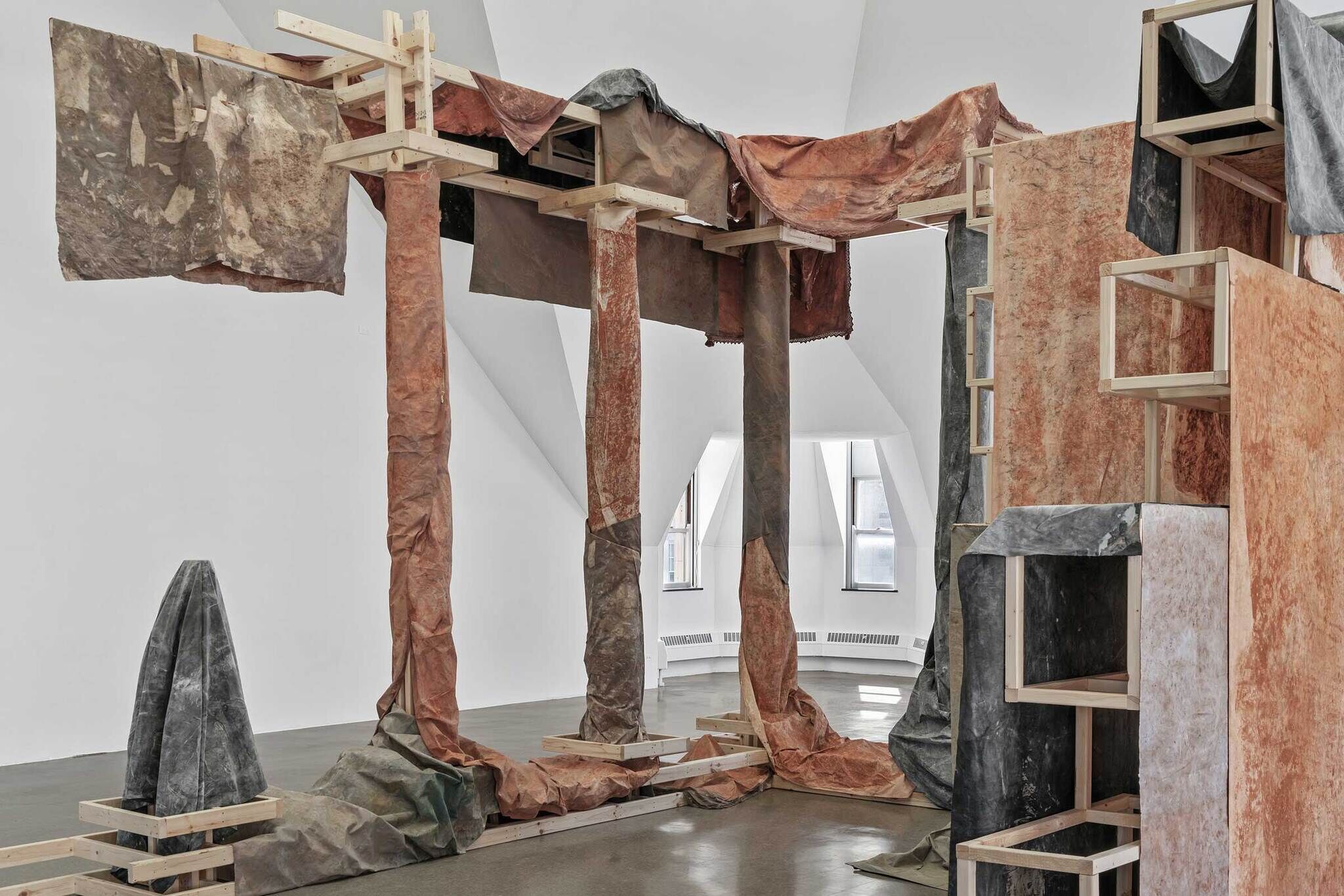 Modern art installation with wooden structures and draped fabrics in a white gallery space.