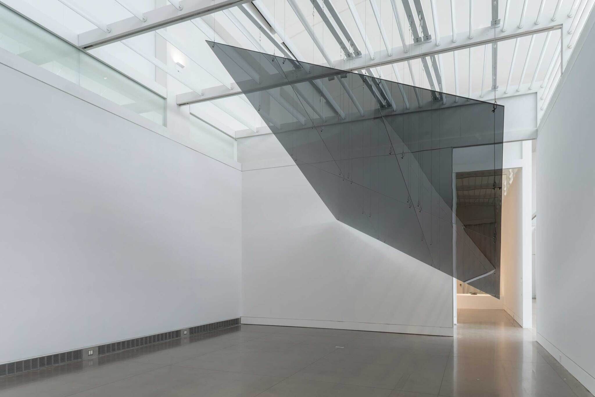 Modern gallery interior with a large, angled, reflective art installation suspended from the ceiling.