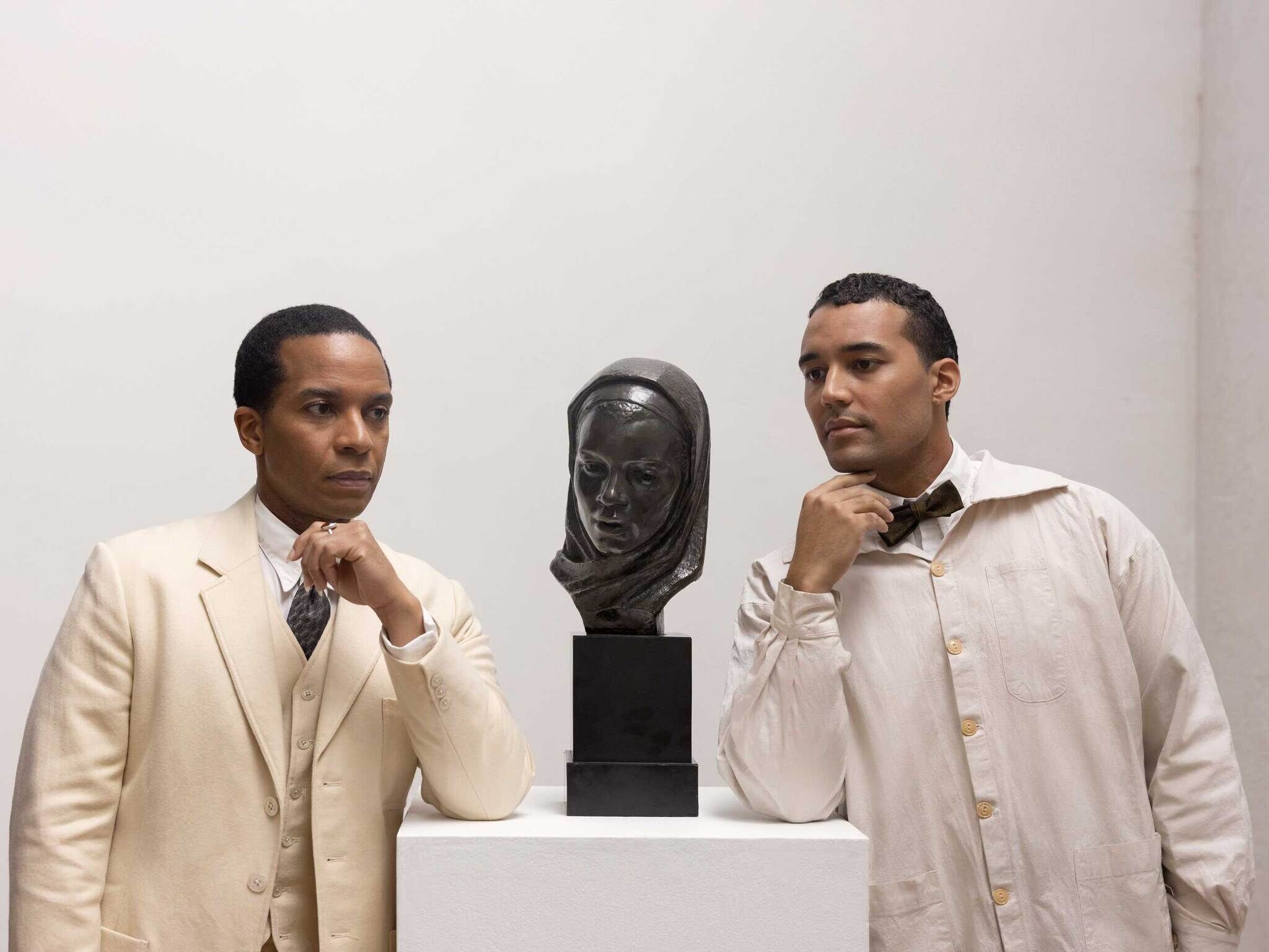 Two men in light suits posing thoughtfully beside a bust sculpture on a pedestal.