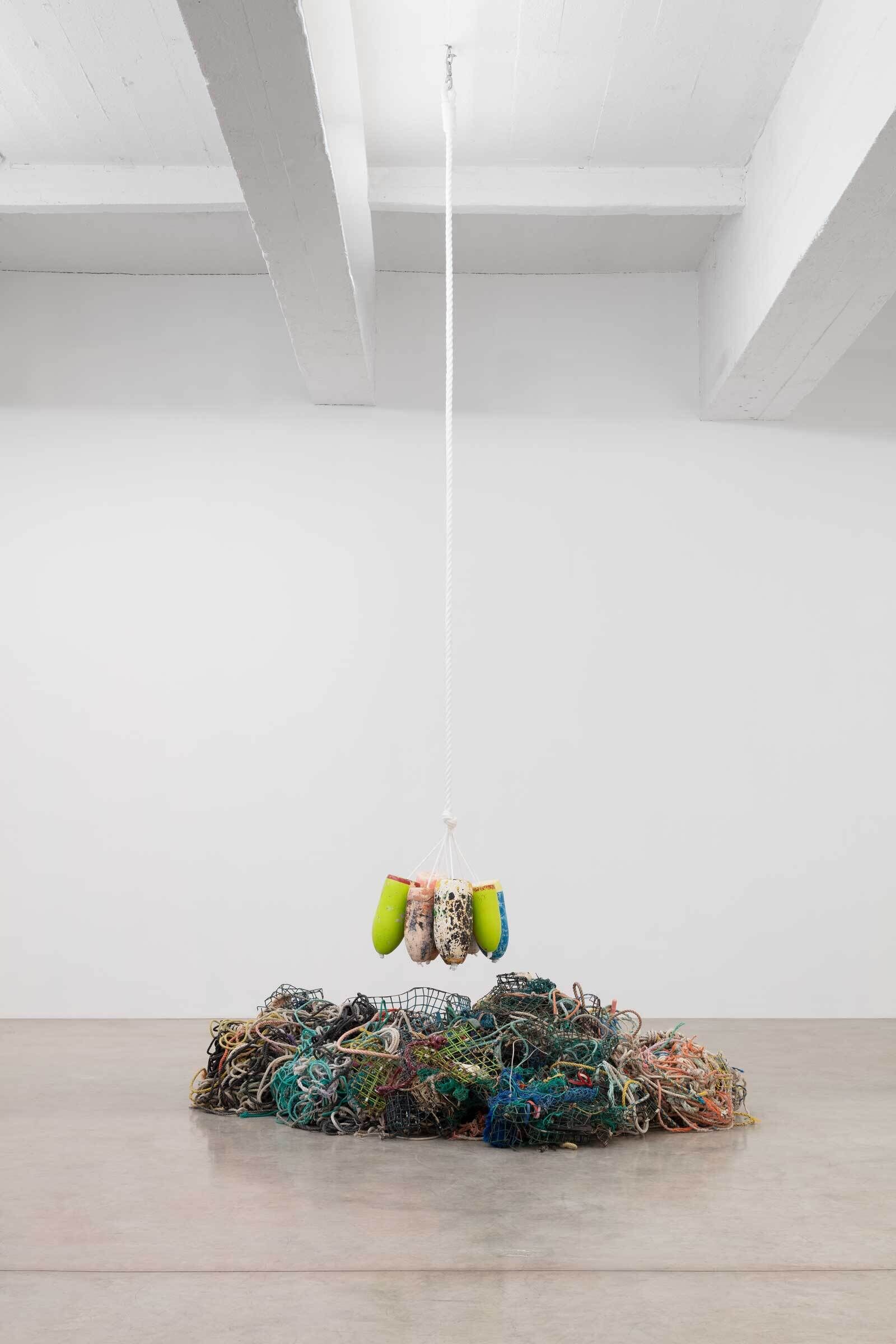 Colorful buoys hanging from a rope above a pile of tangled fishing nets on a gallery floor.
