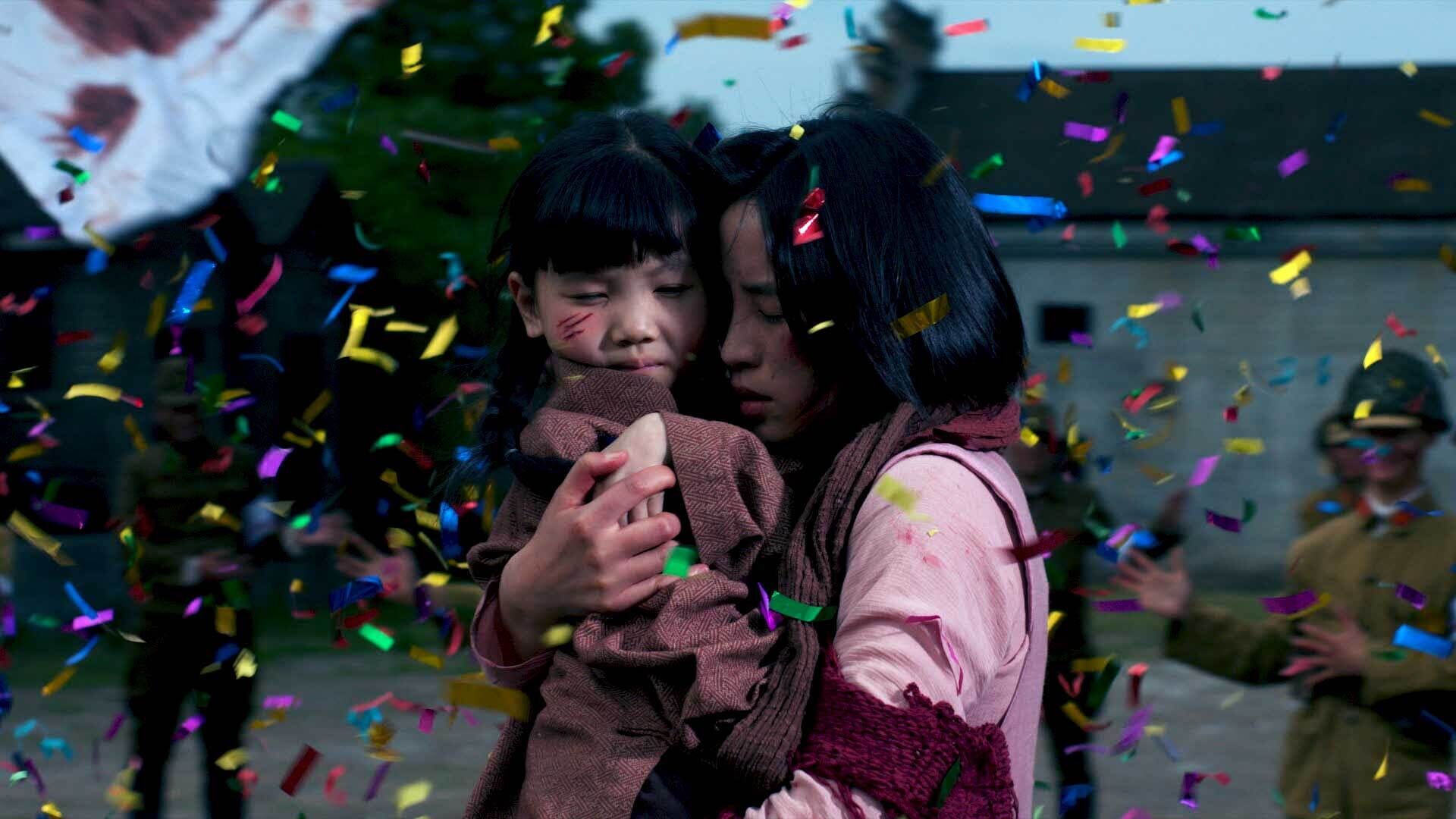 A woman and child embrace amid falling confetti, with one comforting the other who appears distressed with a bloody mark on their face.