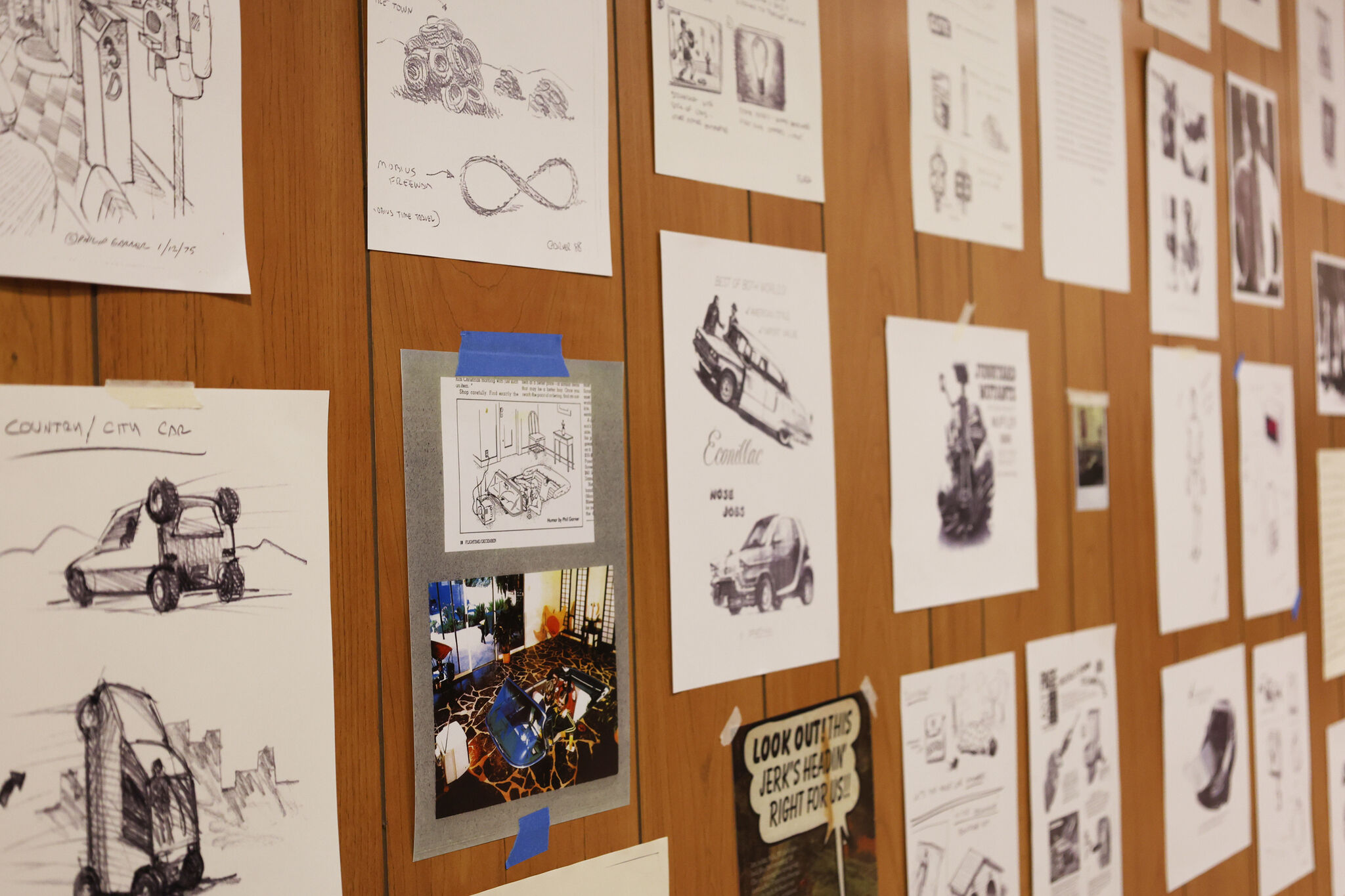 A wood panel wall covered with various sketches and printed images, mostly related to automotive designs and concepts.