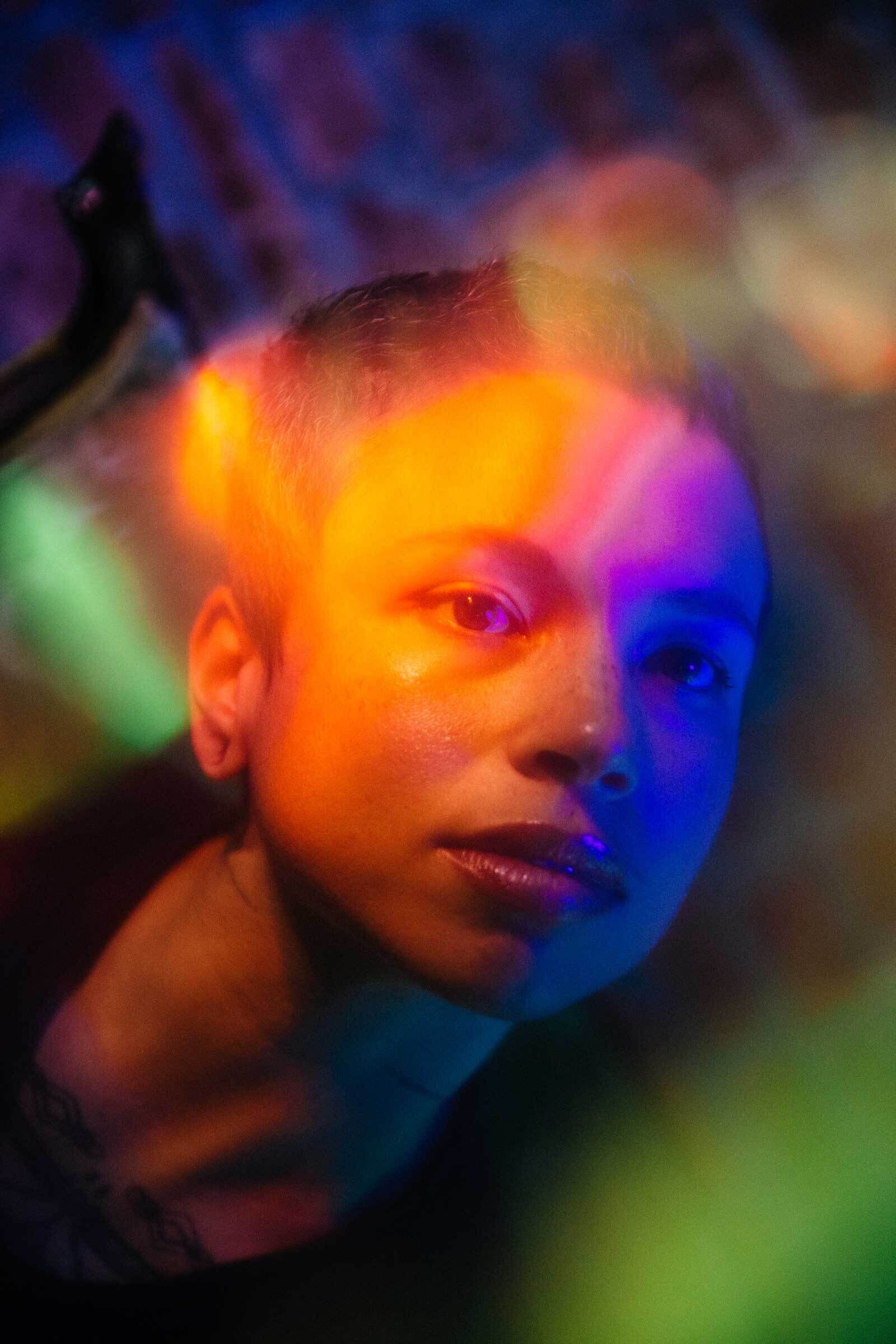 Person with a buzz cut in colorful lighting looking thoughtfully to the side.