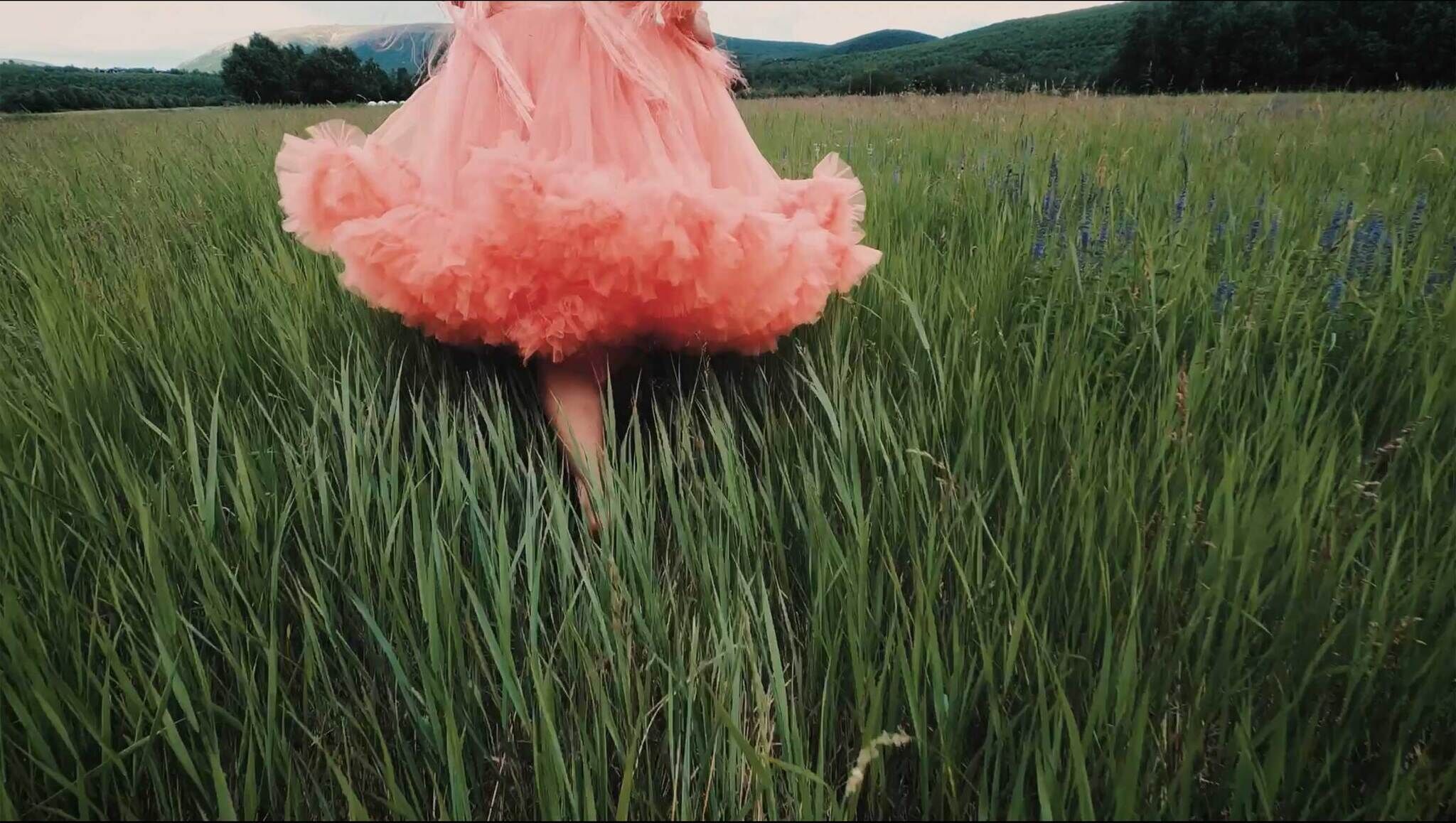 Person in a billowing peach dress mid-twirl in a lush green field with distant hills.