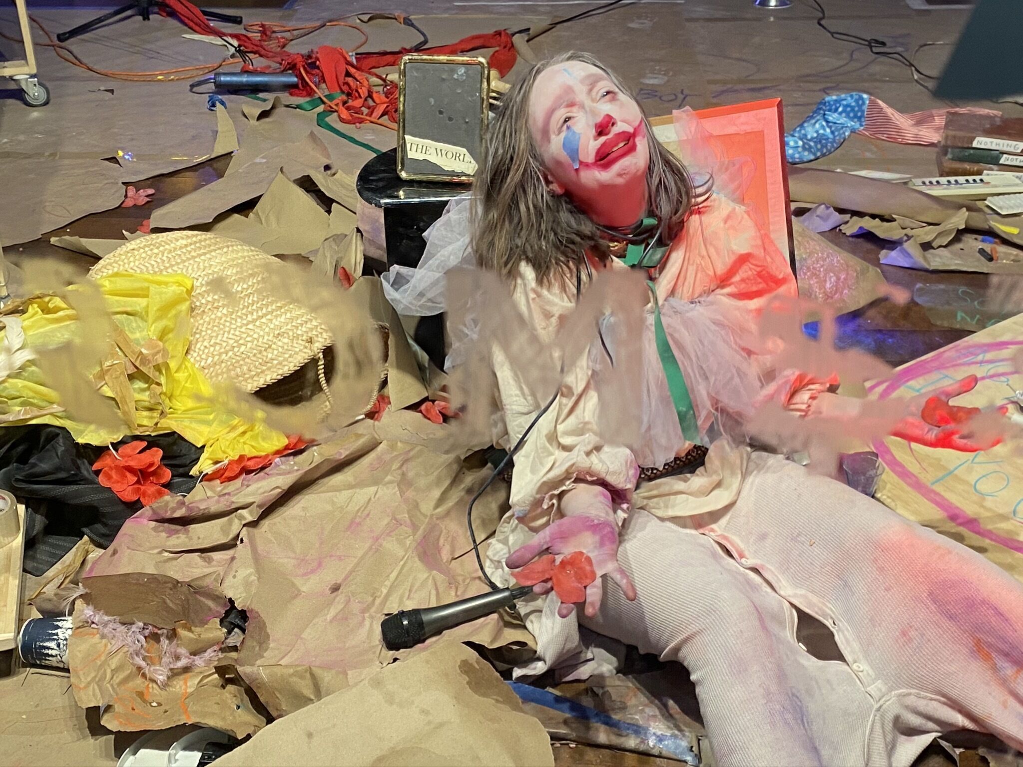Person with clown makeup amidst chaotic scene with scattered props and colorful debris.