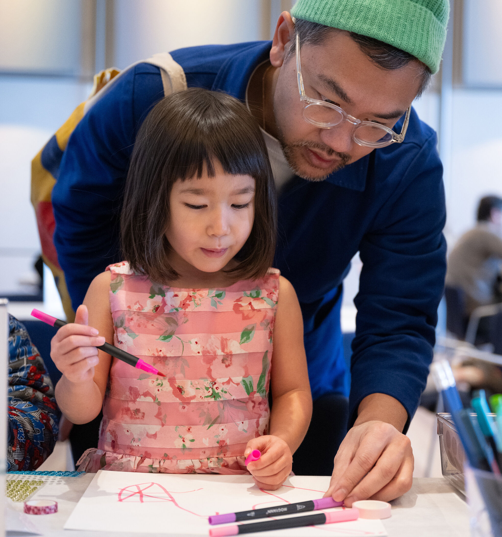 Man in green beanie attentively watches a young girl drawing with markers at a table.