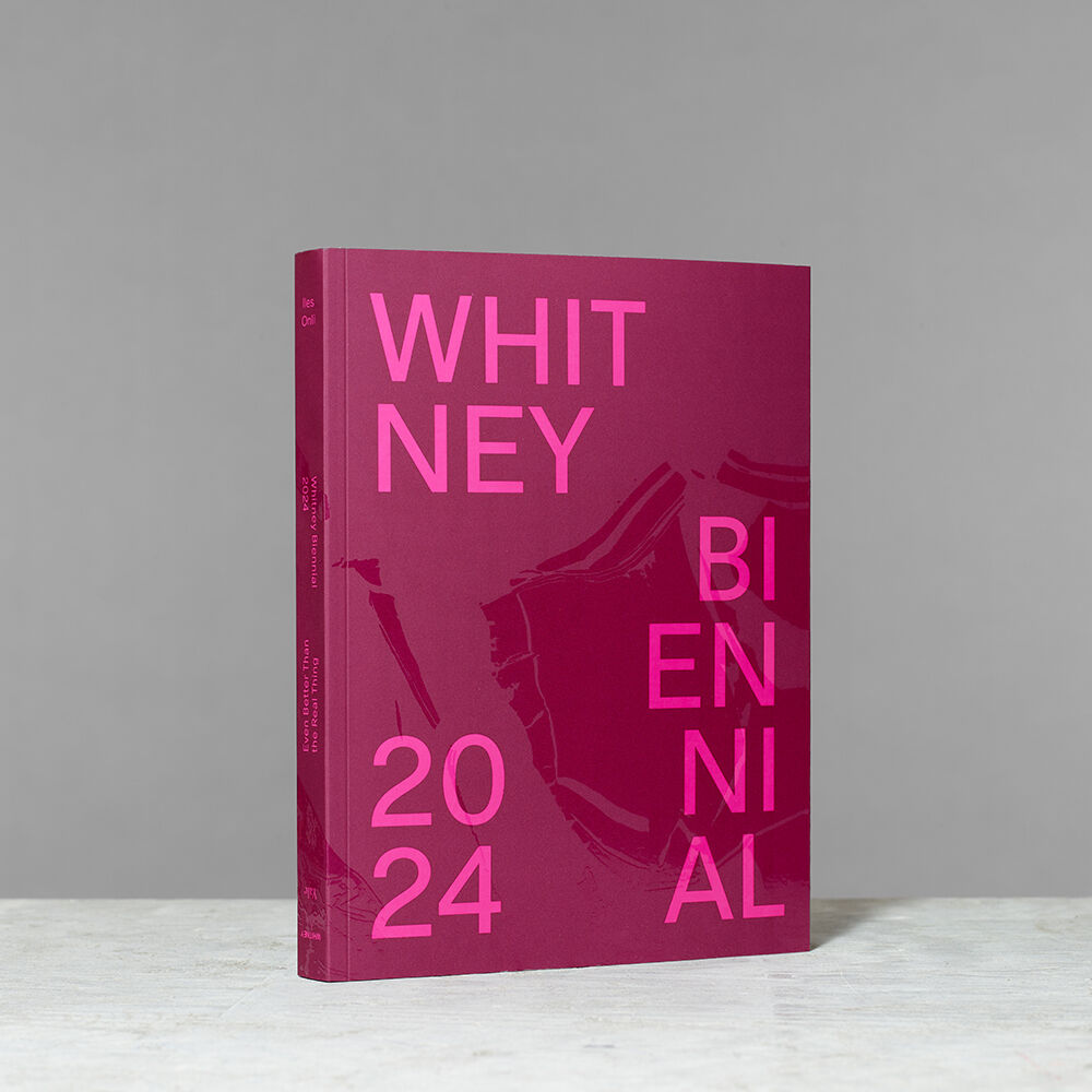 A pink book titled "WHITNEY BIENNIAL 2024" standing upright on a gray surface against a neutral background.
