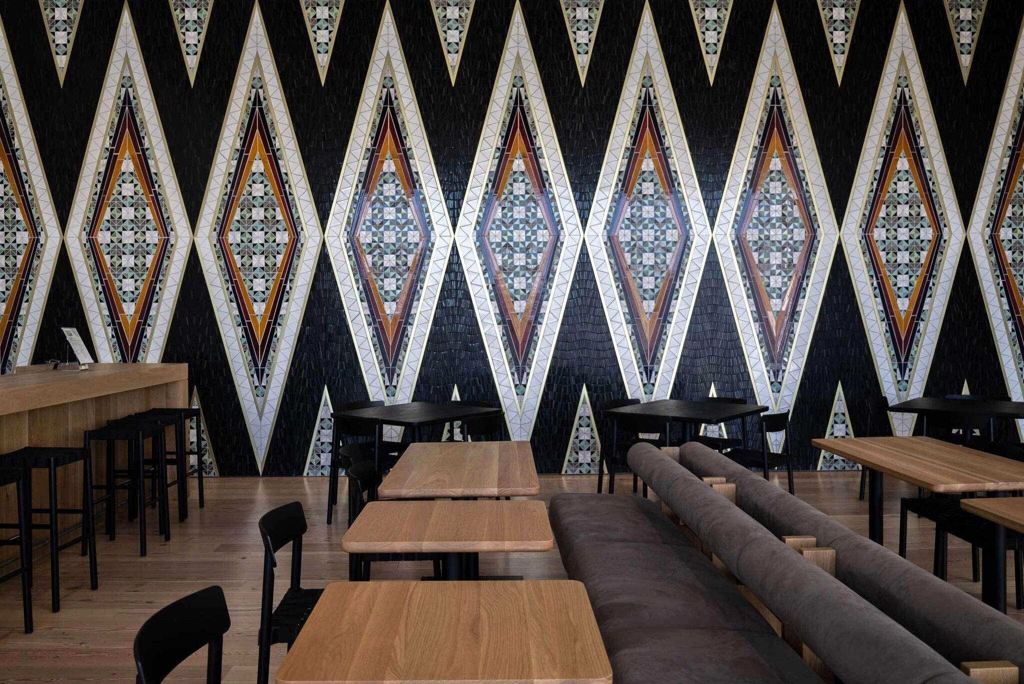 Modern cafe interior with geometric patterned on a mosaic wall, wooden tables, black chairs, and a long cushioned bench.