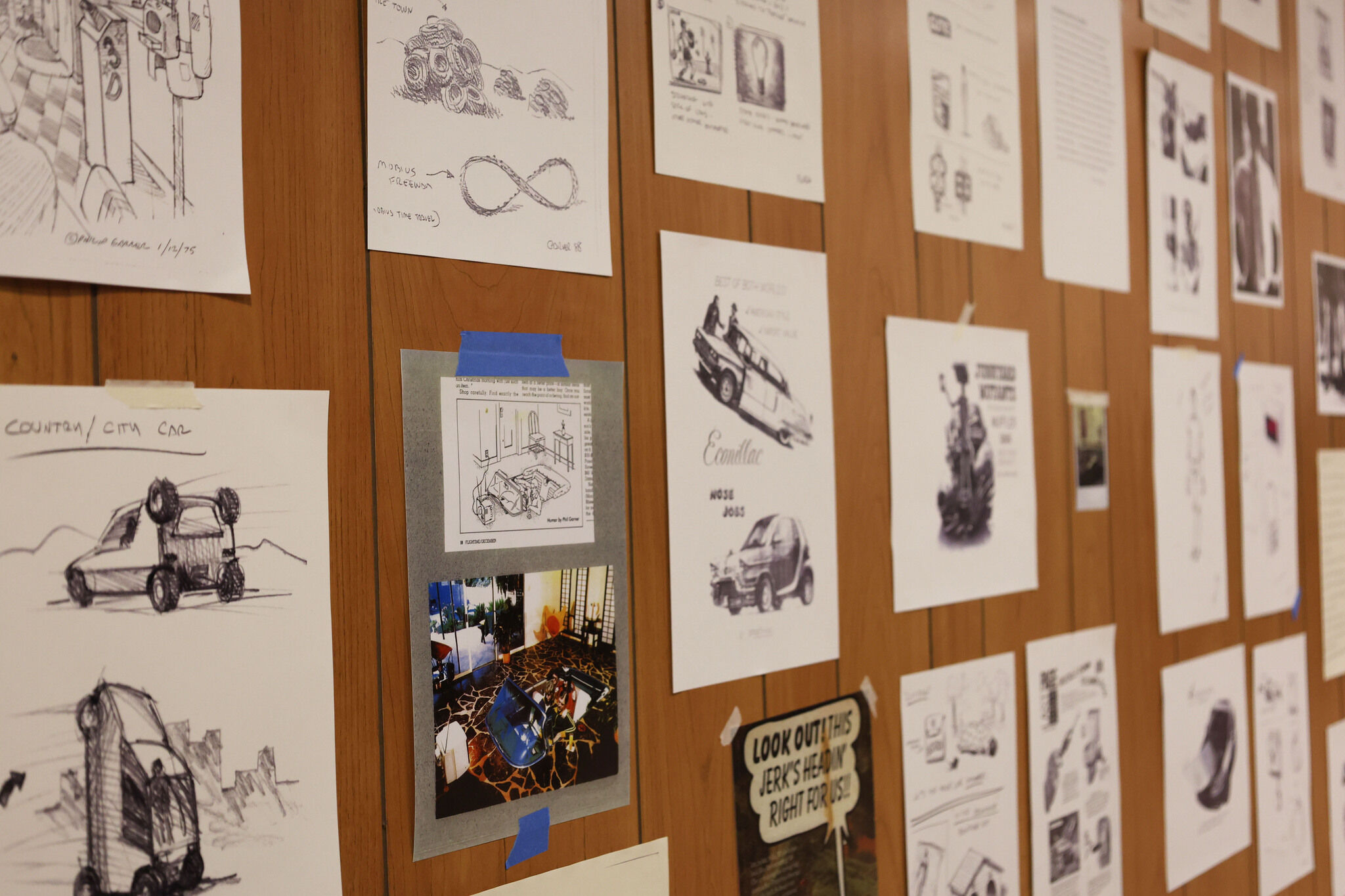A bulletin board covered with various sketches and printed illustrations, some related to cars and design concepts.