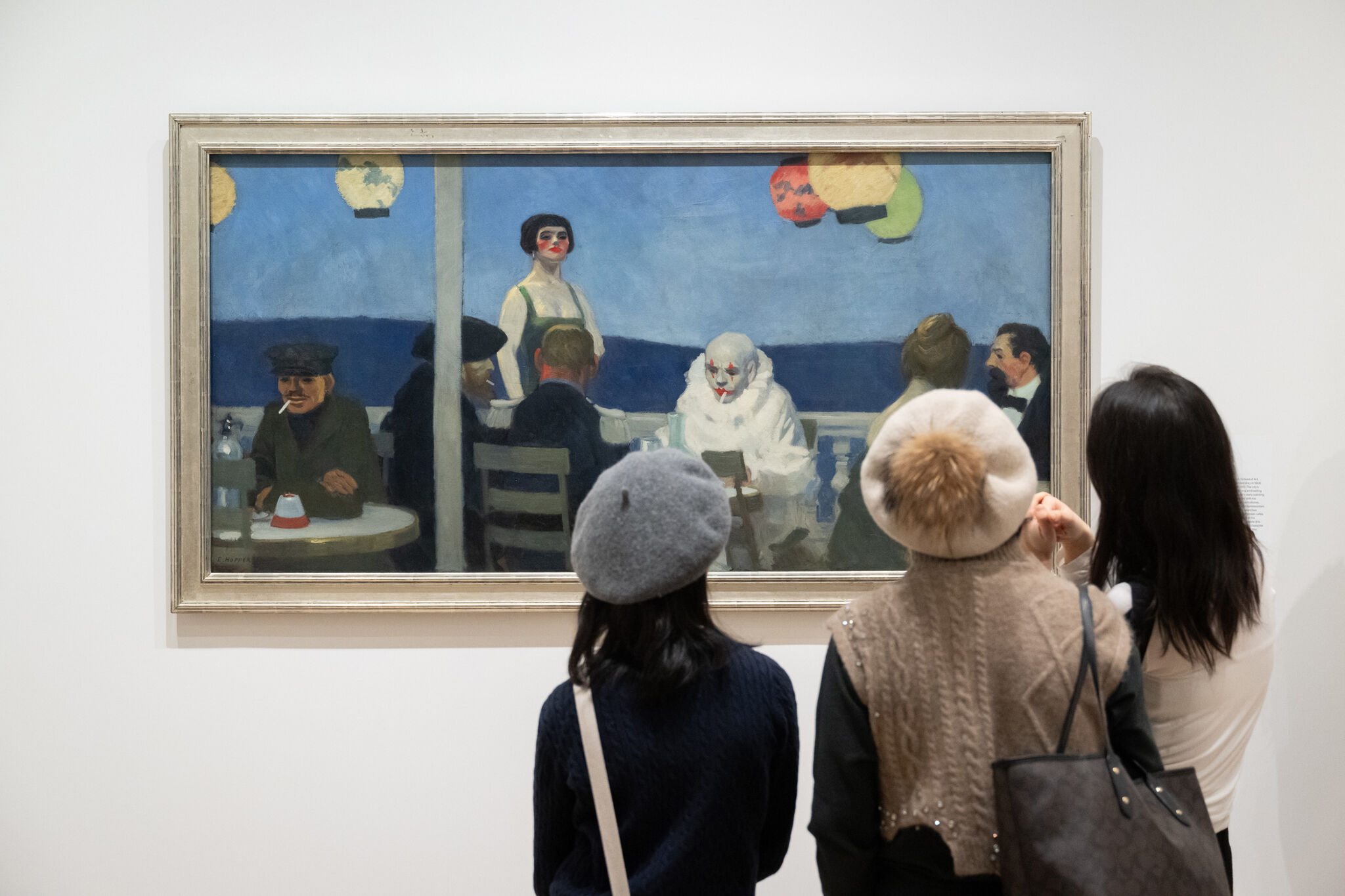 Visitors at a gallery observing a painting of people dining at a night-time café setting.