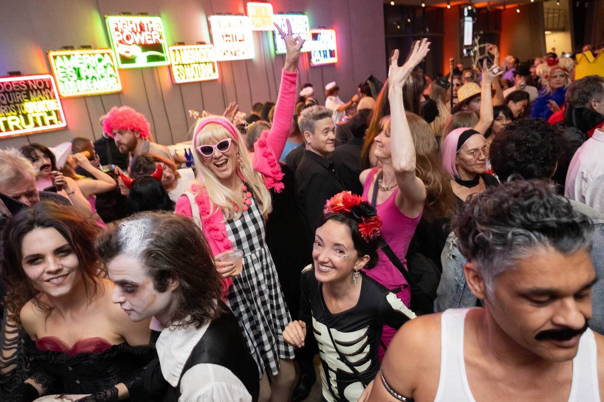 Crowd of people dancing in front of a bar int he Whitney Museum Lobby. Guests in costume with pink hair and retro glasses featured at center.