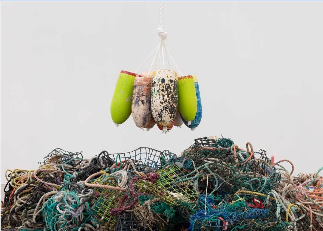 Worn buoys in speckled black and white or bright green and blue hang on a knotted white rope above a jumbled mass of fishing nets, ropes, wires, and strings. 