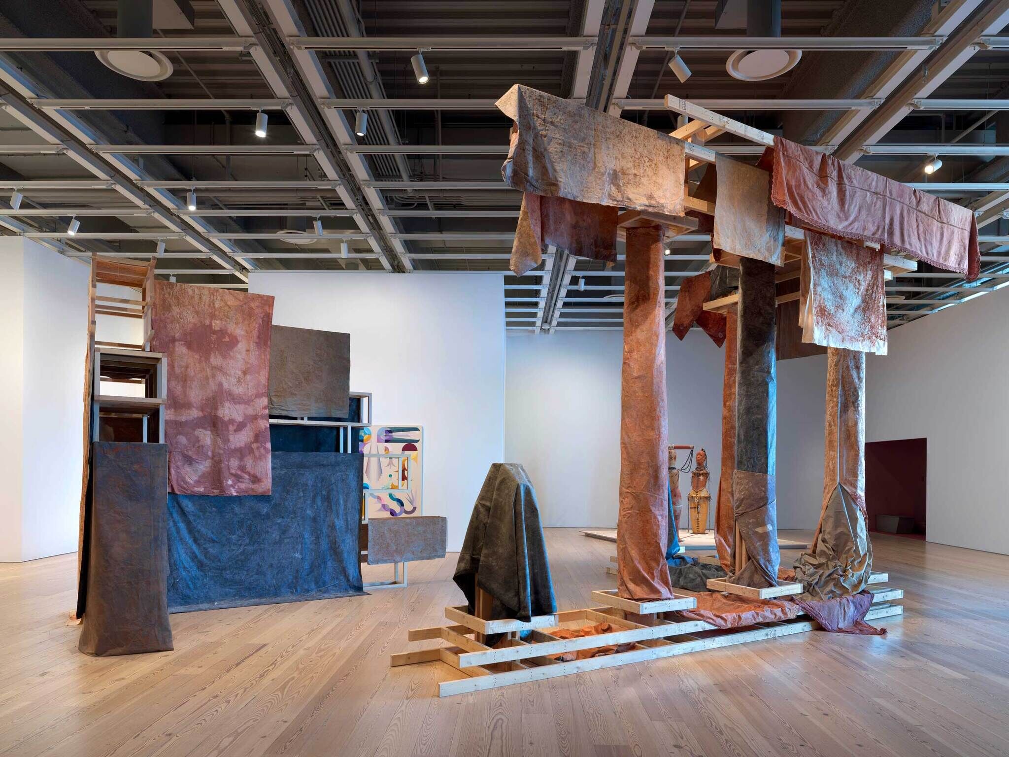 Modern art installation with abstract wooden structures and draped fabrics in a gallery setting.