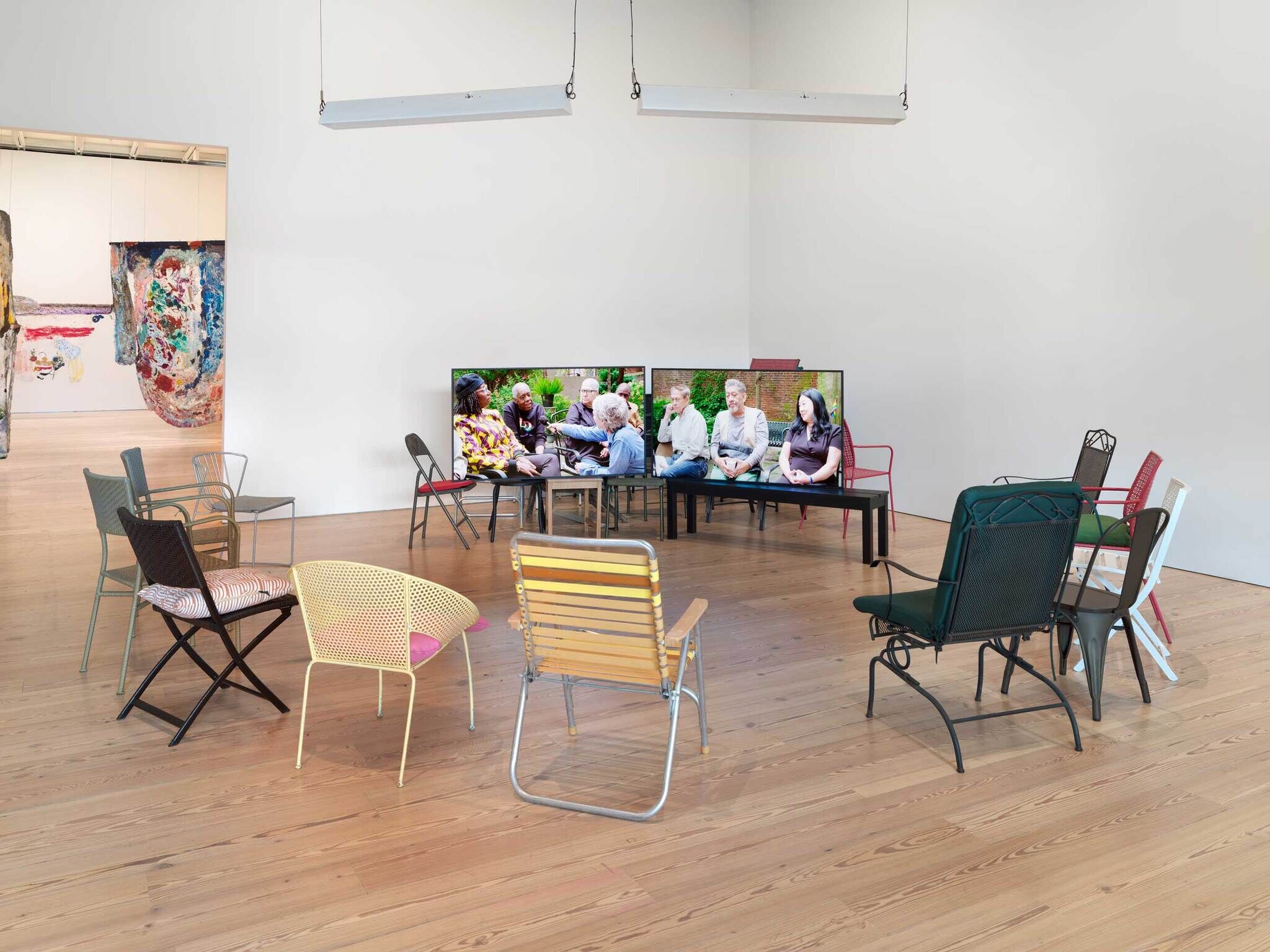 Art gallery interior with colorful chairs scattered around and a large video screen displaying people in conversation.