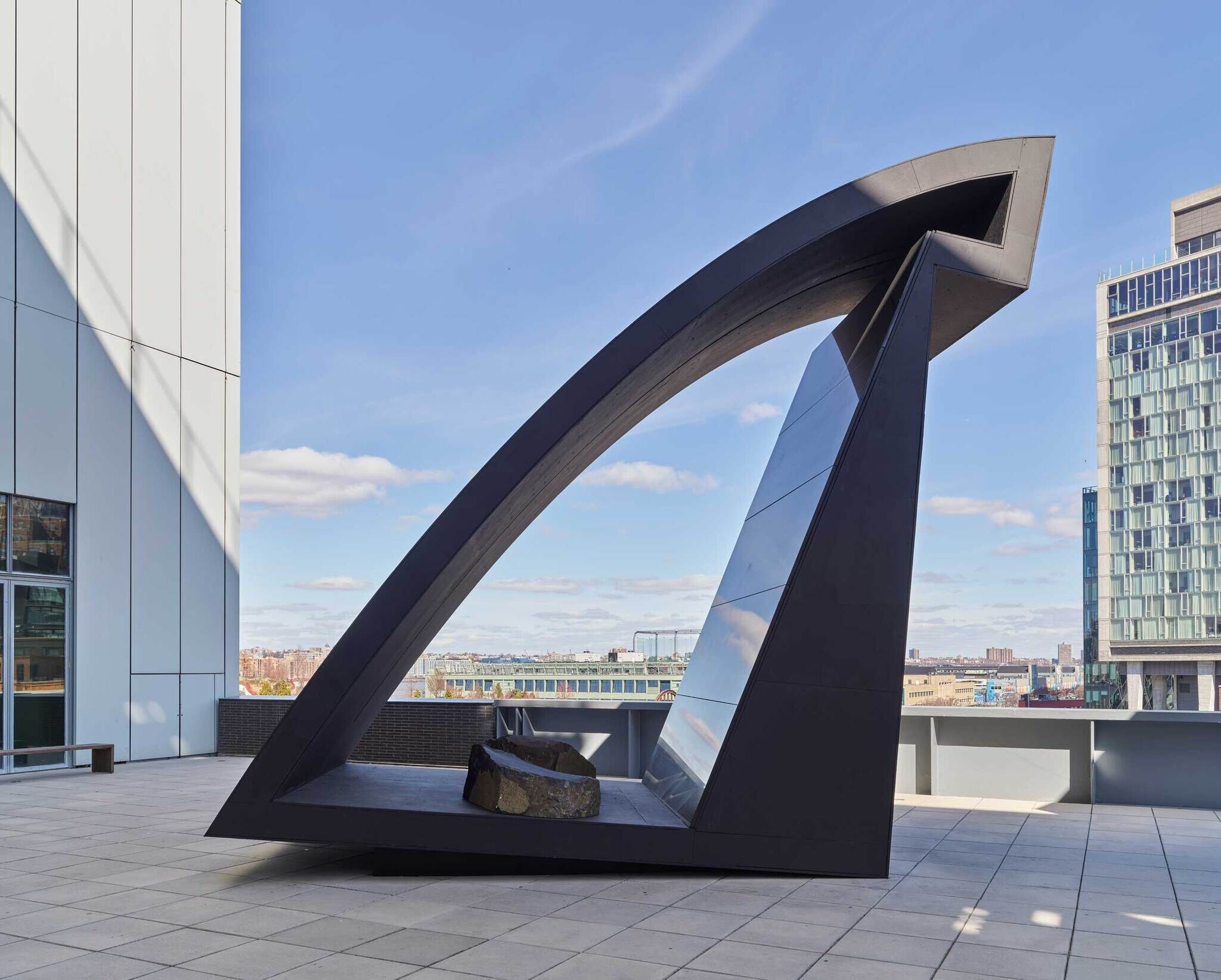 Modern outdoor sculpture resembling a stylized triangular portal, with city skyline and blue sky in the background.