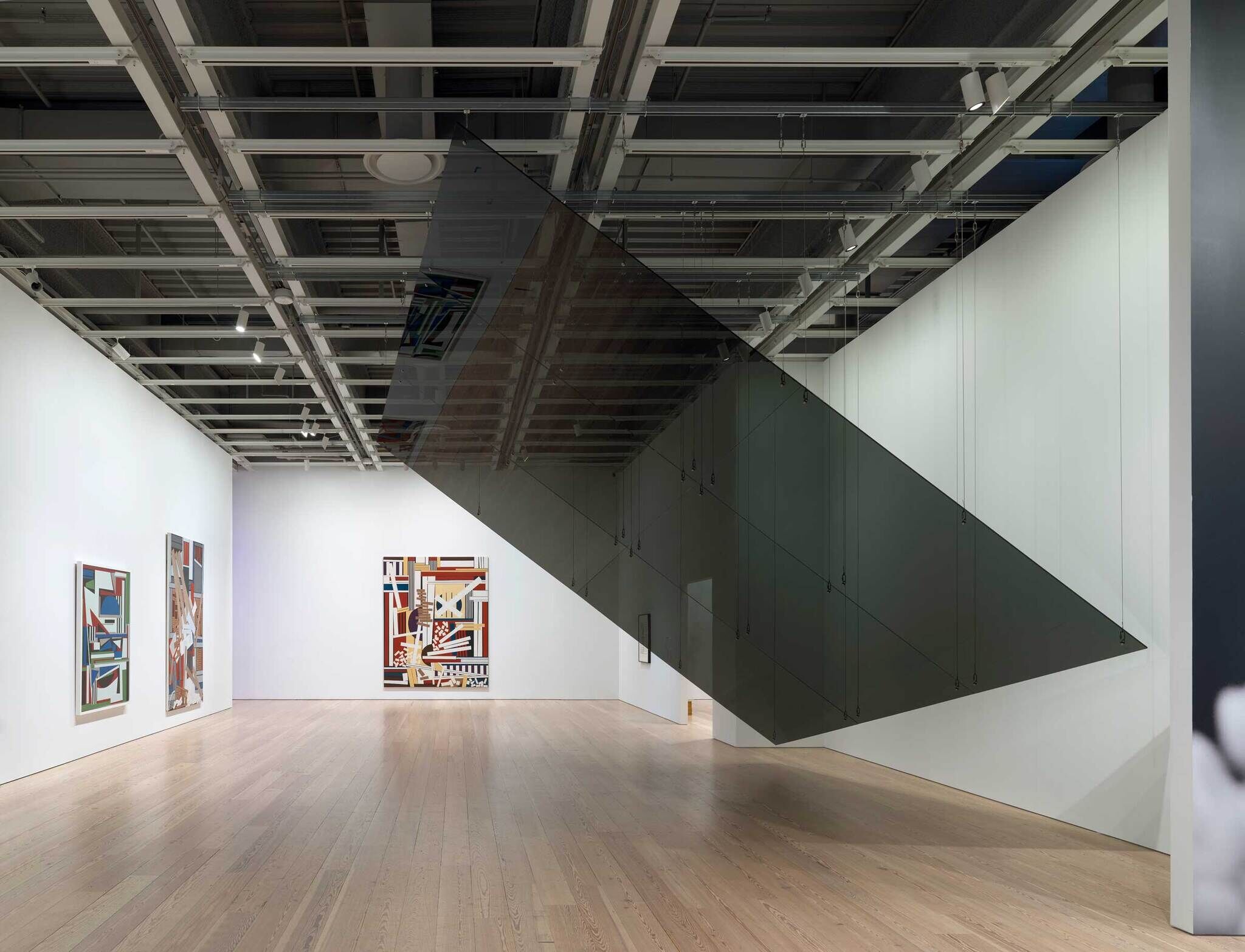 Modern art gallery interior with abstract paintings on white walls and a large, angled mirror installation reflecting the space.