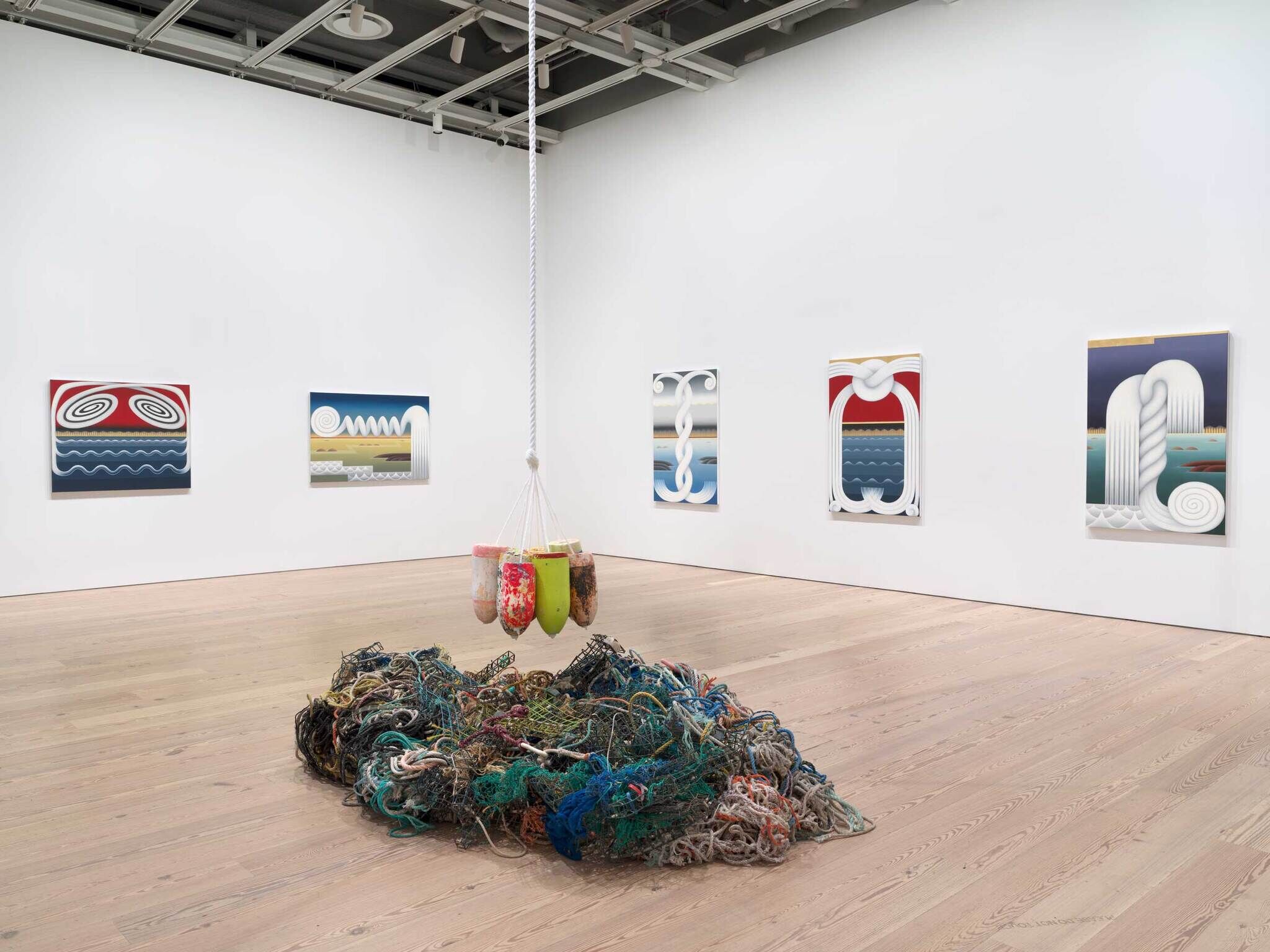 Modern art gallery with colorful abstract paintings on white walls and a pile of tangled fishing nets on the floor.