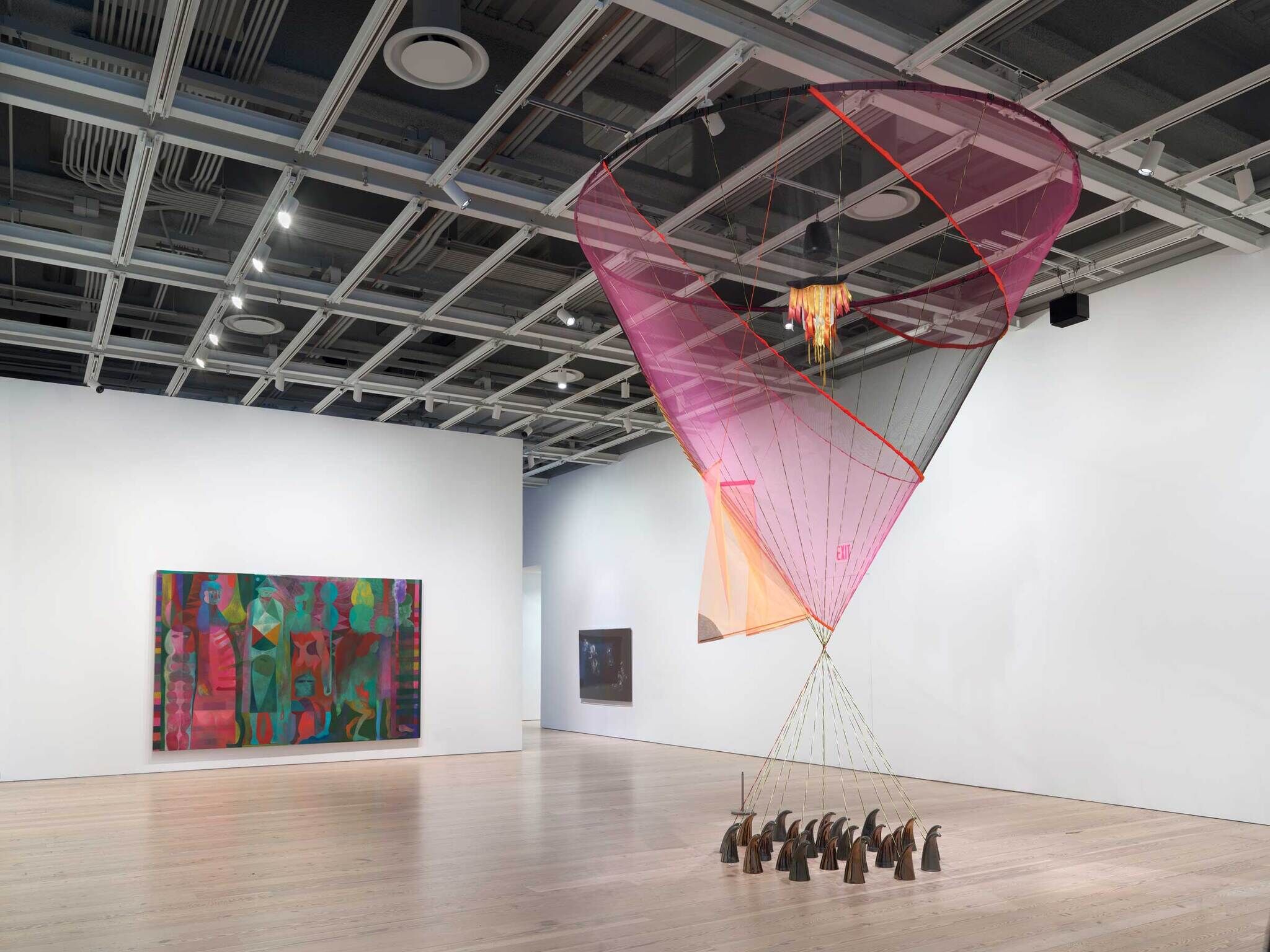 A large, colorful suspended sculpture resembling an upside down teepee dominates an art gallery with paintings on the walls.