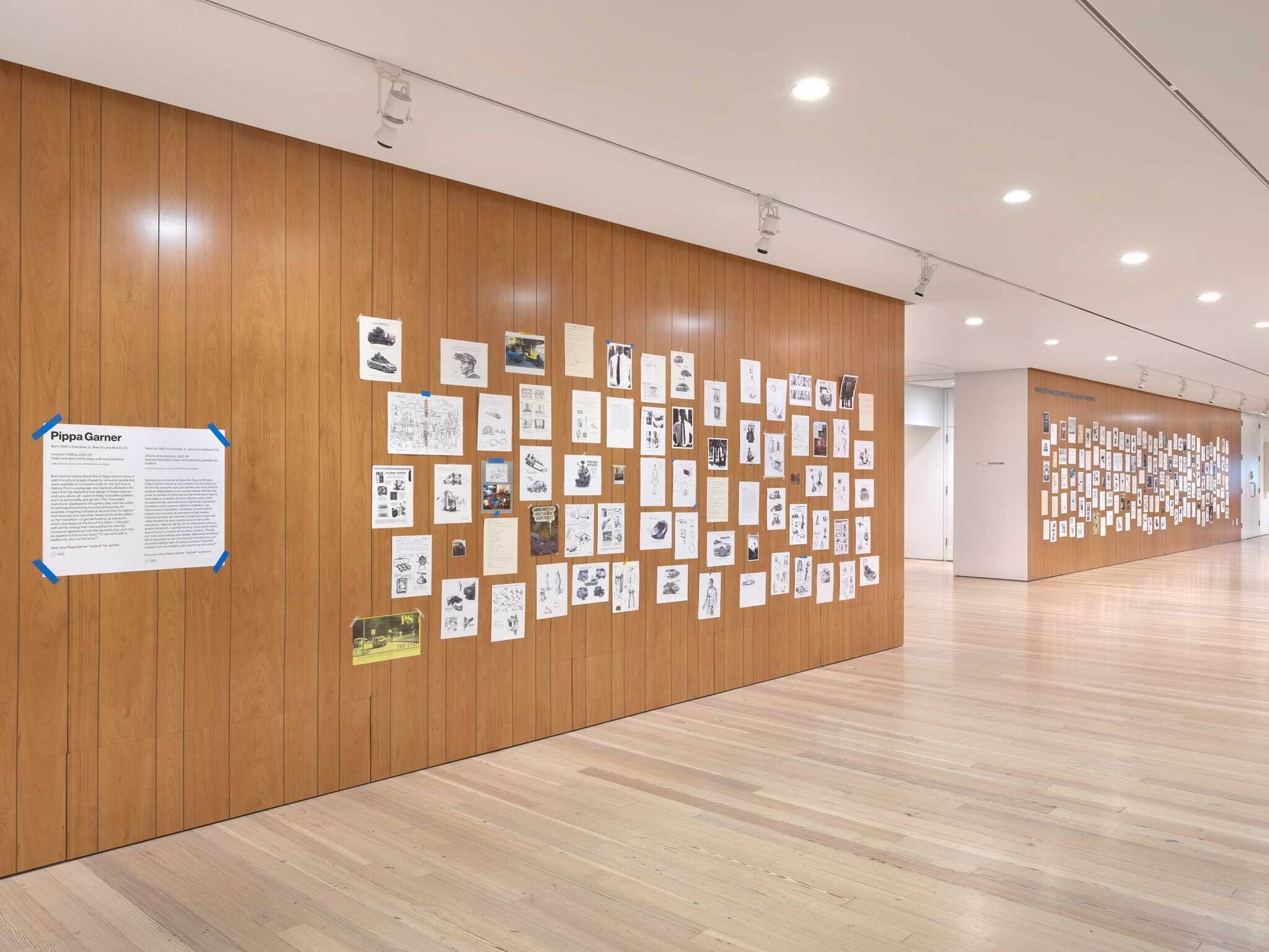 A gallery interior with a wooden wall displaying numerous sketches and images, accompanied by an informational poster on the left.
