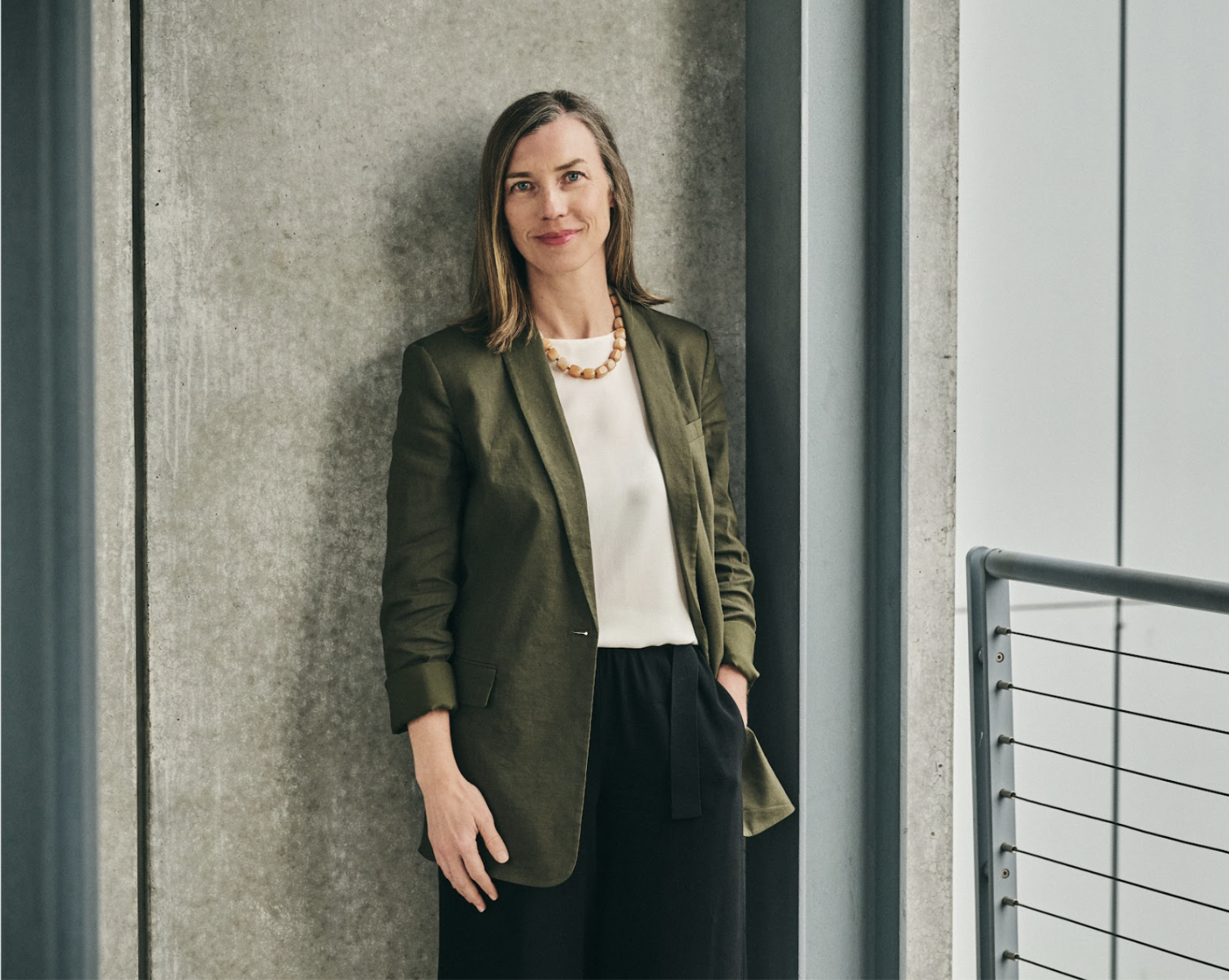 Professional woman in a green blazer and black pants standing by a concrete wall with a relaxed posture.
