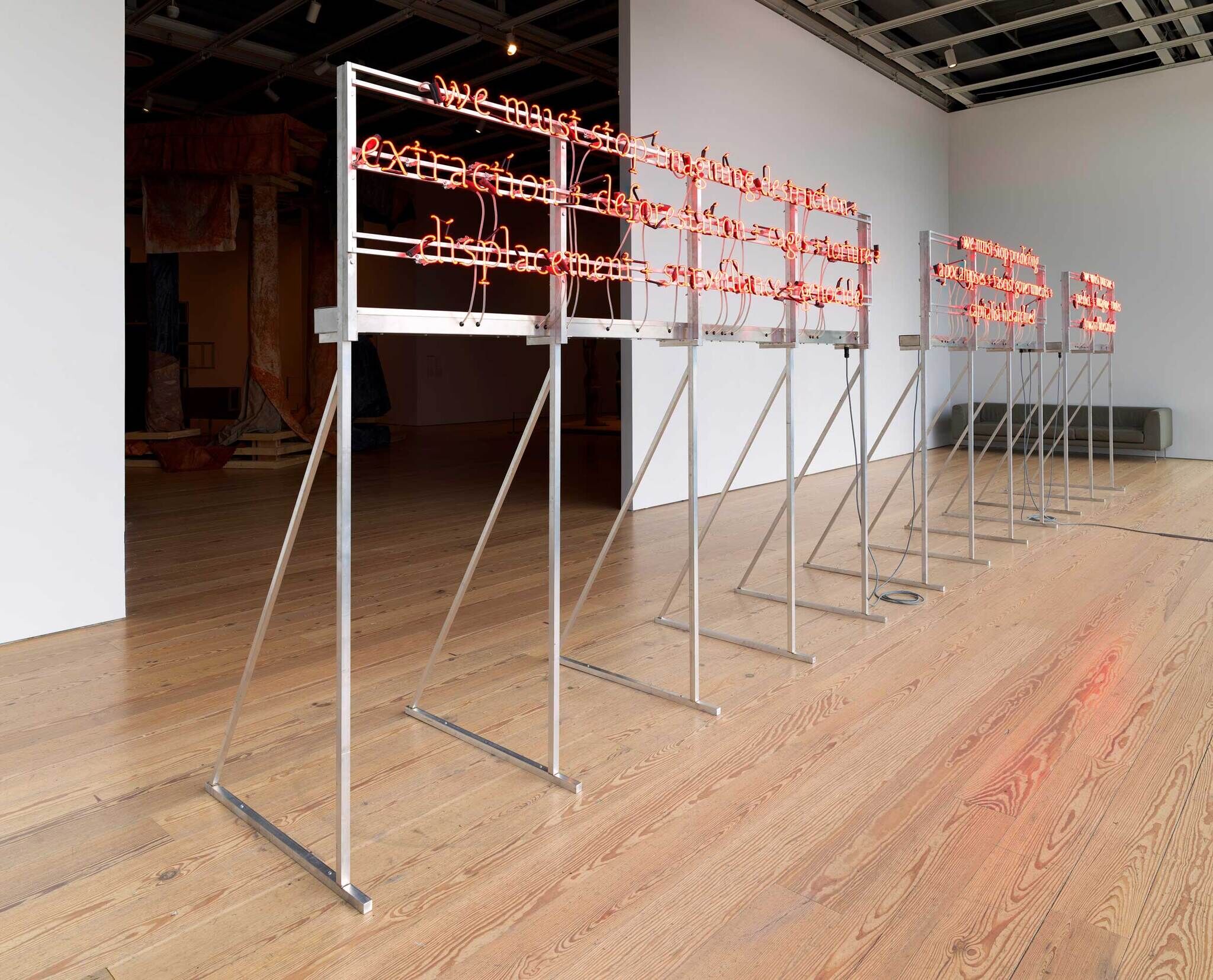 Neon light installation with text on metal stands in an art gallery, wooden floors, and abstract art in the background.