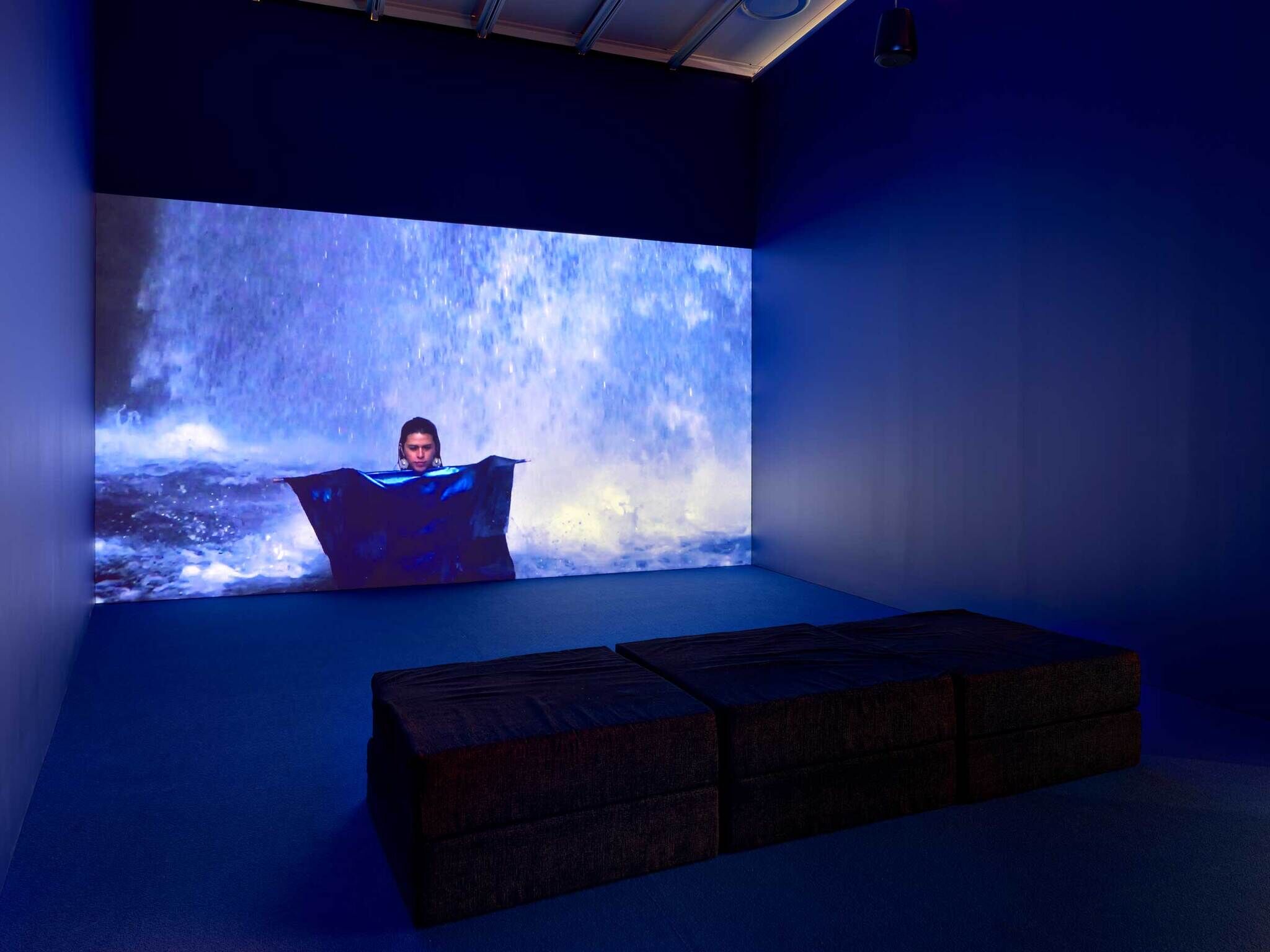 A person emerges from a blue tarp in front of a large video screen displaying a waterfall, with dark cushions on the floor.
