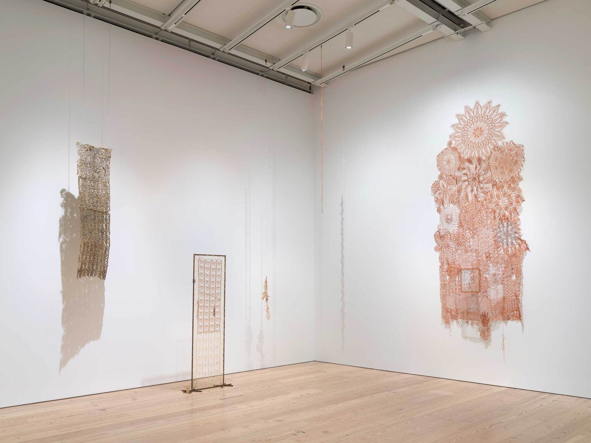 Intricate lace-like textile art pieces displayed in a bright gallery space, hanging from the ceiling and standing on the floor.