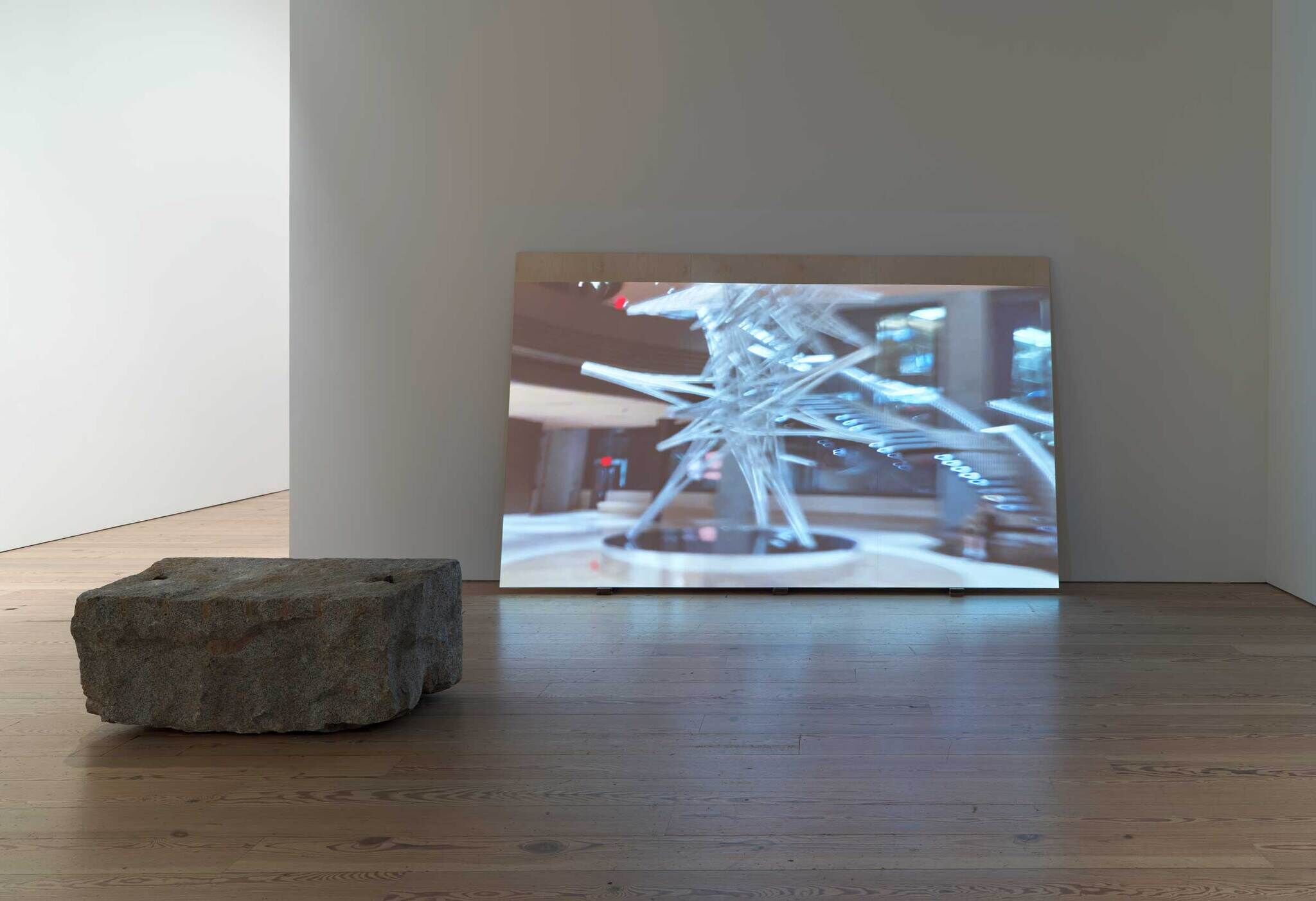 A large stone block on a wooden floor with a blurred photo of a sculpture leaning against a wall in a gallery.