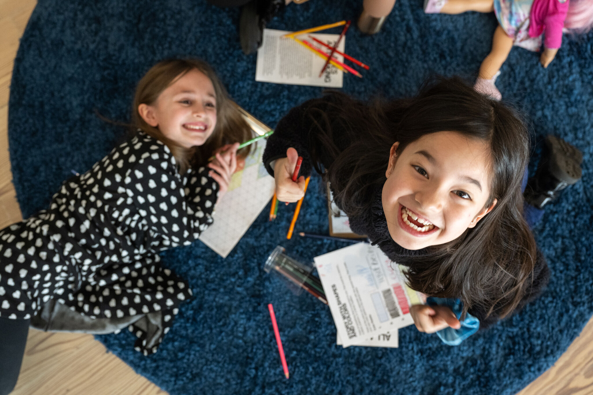 Two girls lying on a blue rug, smiling at the camera with coloring supplies scattered around them.