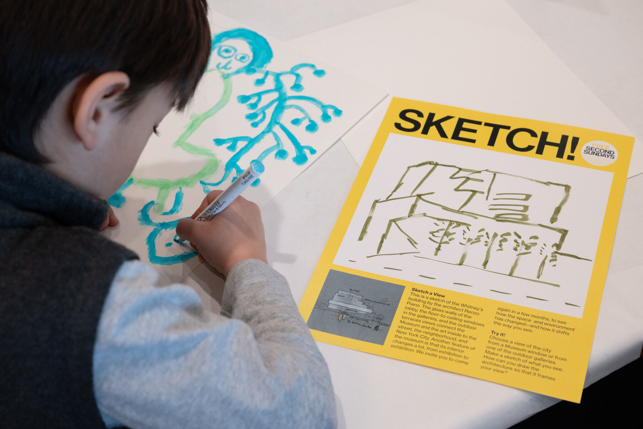 A child draws on paper next to a "SKETCH" instruction sheet, part of an educational activity.