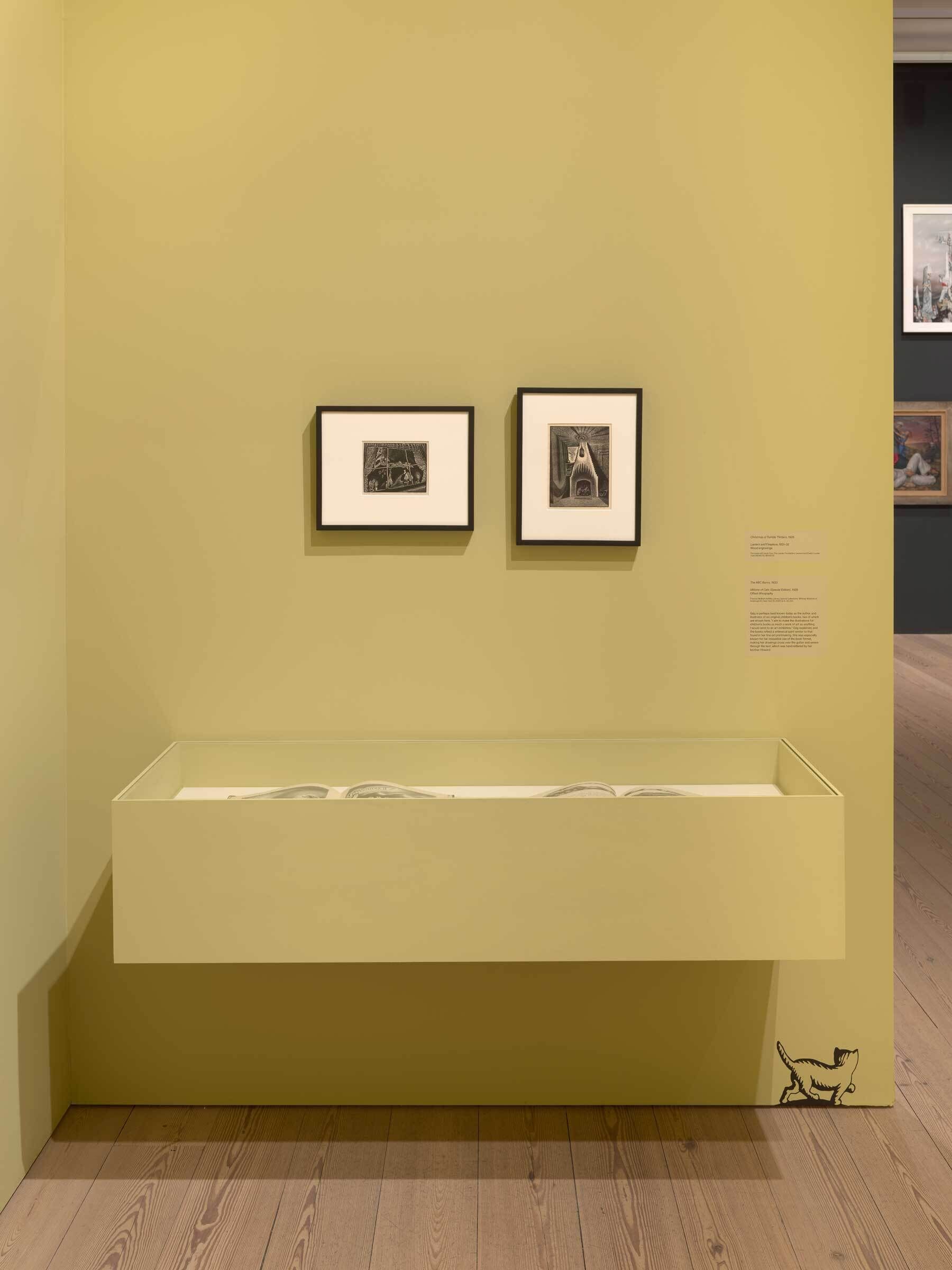 Two framed artworks on a mustard-colored gallery wall above a white bench with a small cat illustration near the floor.