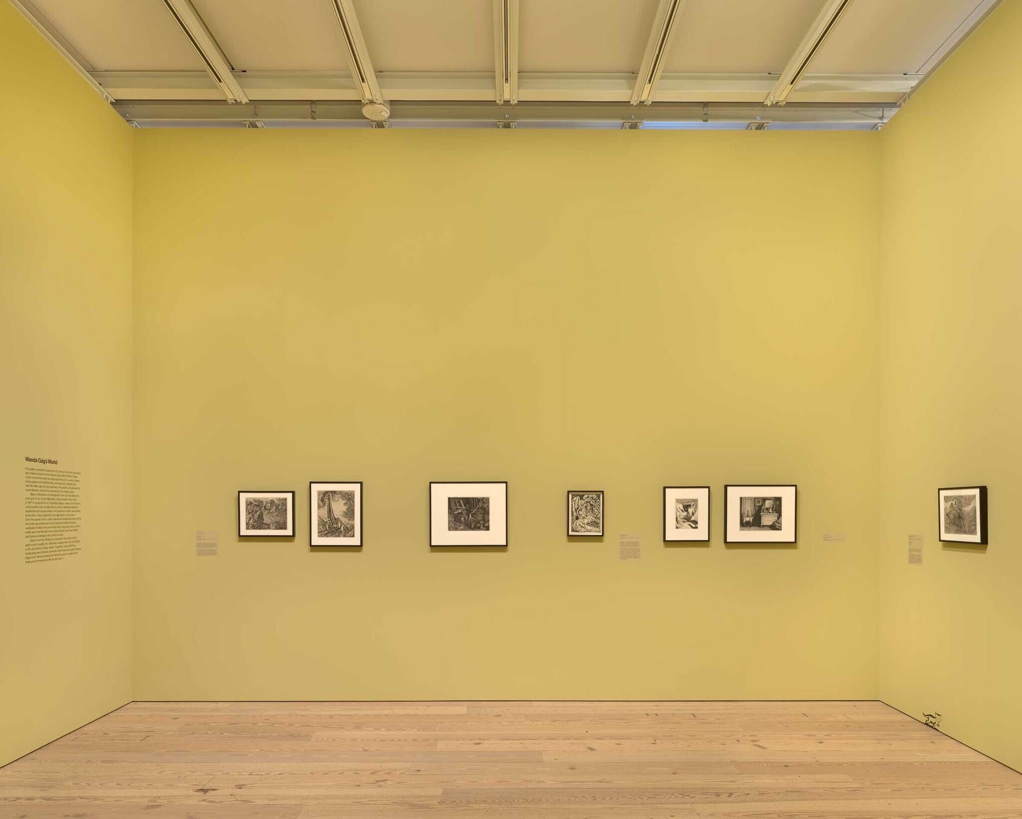 A bright gallery room with yellow walls displaying framed black and white artwork, wooden floors, and track lighting.