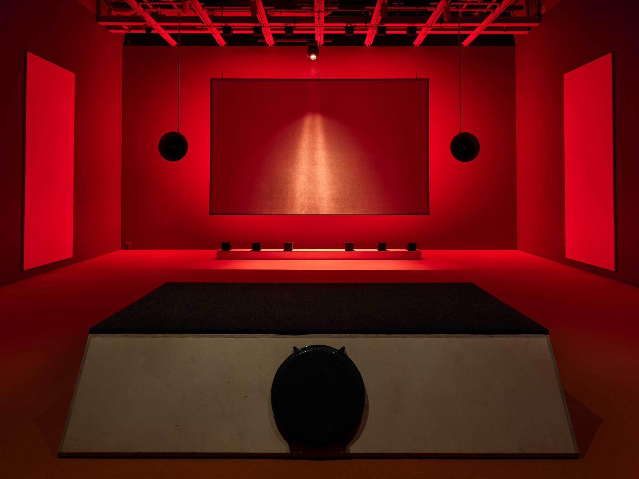 A dimly lit red room with a large screen, speakers, and a stage, evoking a moody cinematic atmosphere.