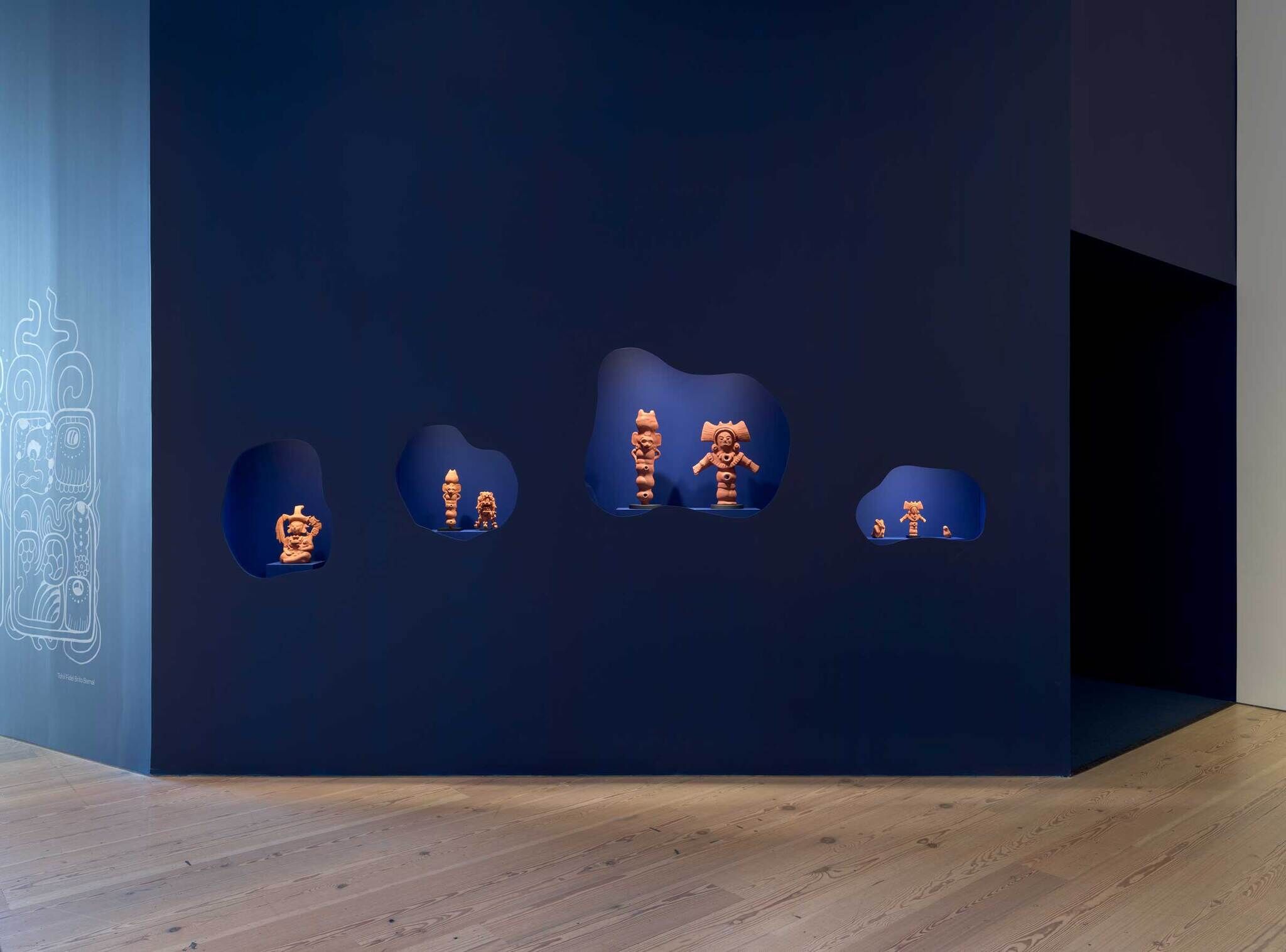 A dark blue gallery wall with irregularly shaped cutouts displaying terracotta sculptures, wooden floor, and a light blue wall with a design on the left.