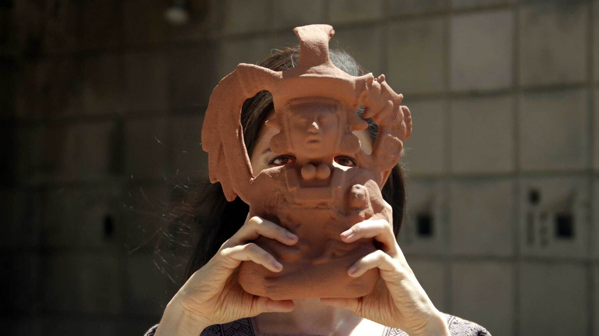A figure with brown hair holds a terracotta Pre-Columbian figurine in front of her face. Her eyes gaze through the gaps in the figurine's intricate clay headdress.