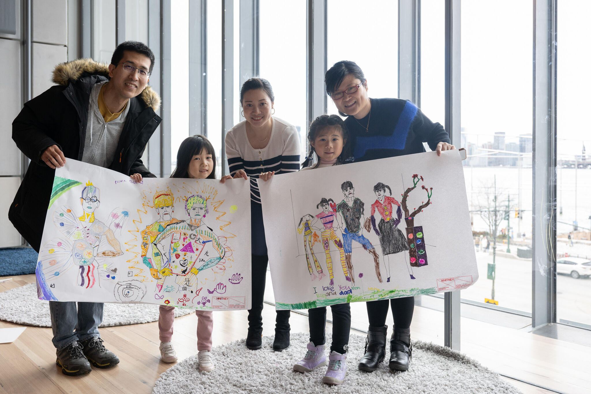 Family proudly displays children's colorful drawings in a bright room with large windows overlooking a cityscape.