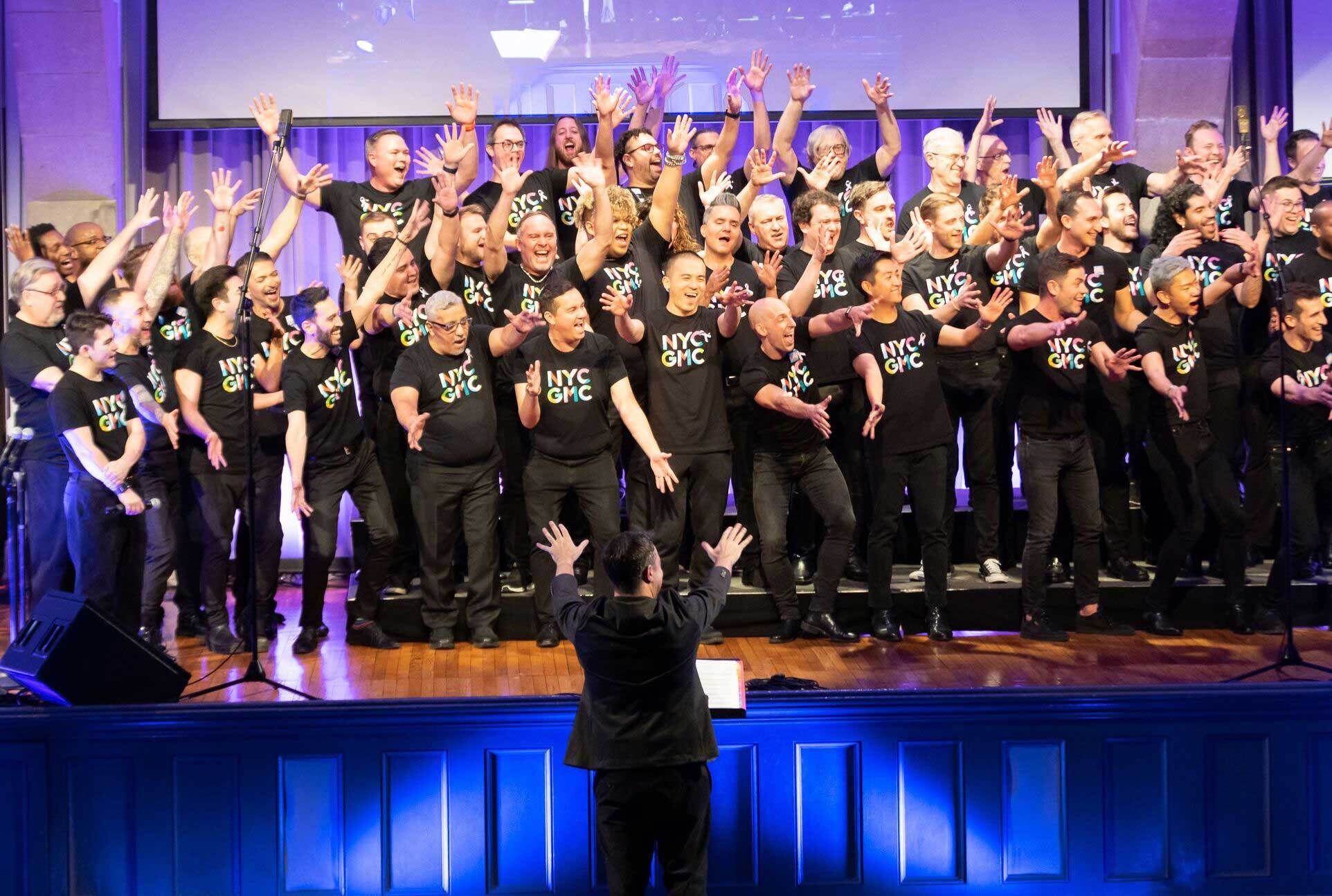 A choir on stage with raised hands, joyfully singing, led by a conductor in front.