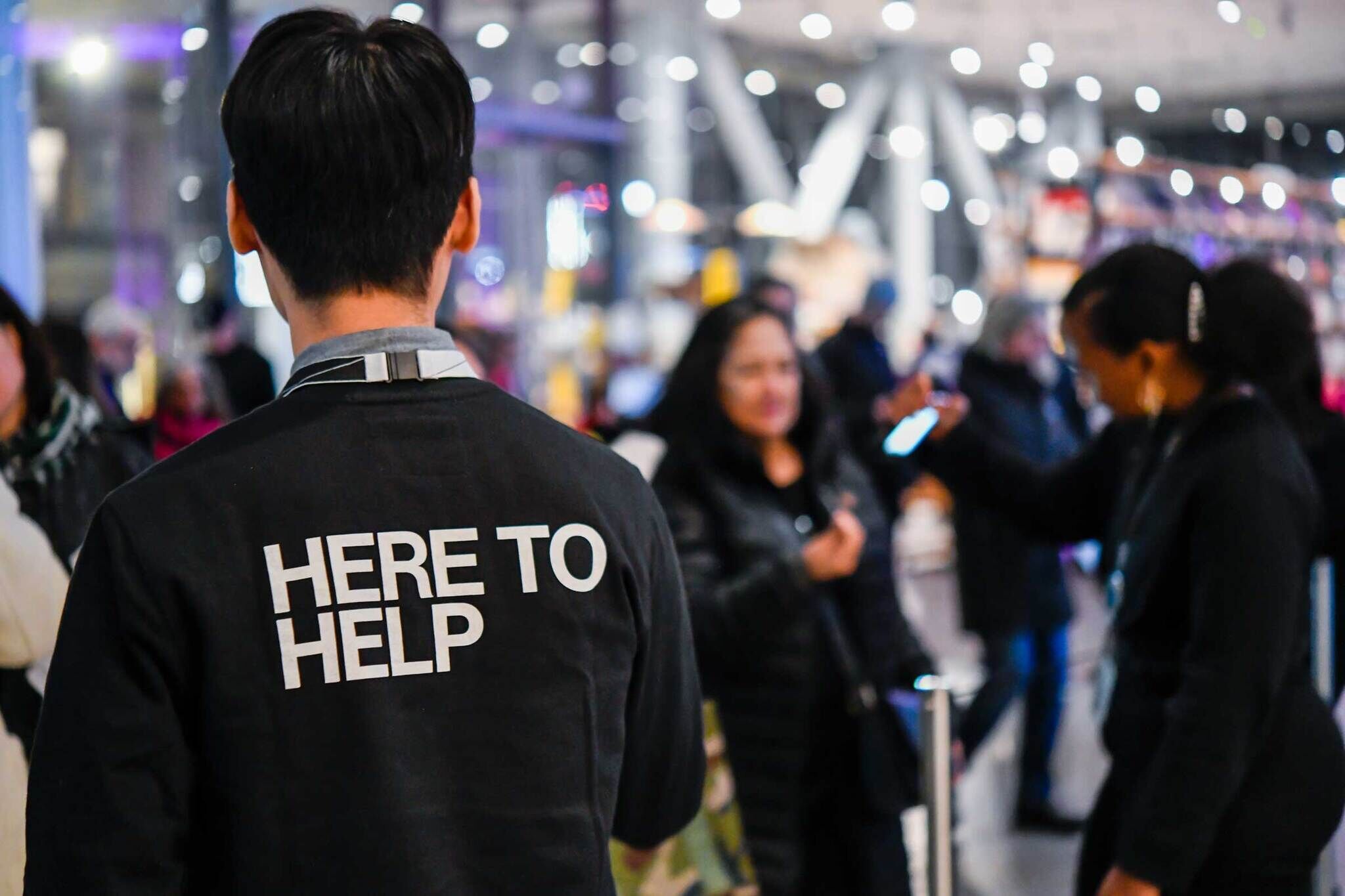 Staff member with "HERE TO HELP" on back of jacket facing a blurred crowd in a busy indoor setting.