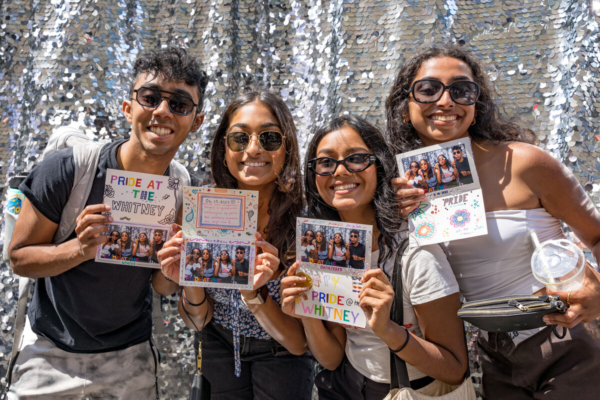 Four smiling friends holding up event flyers in front of a shimmering silver backdrop.