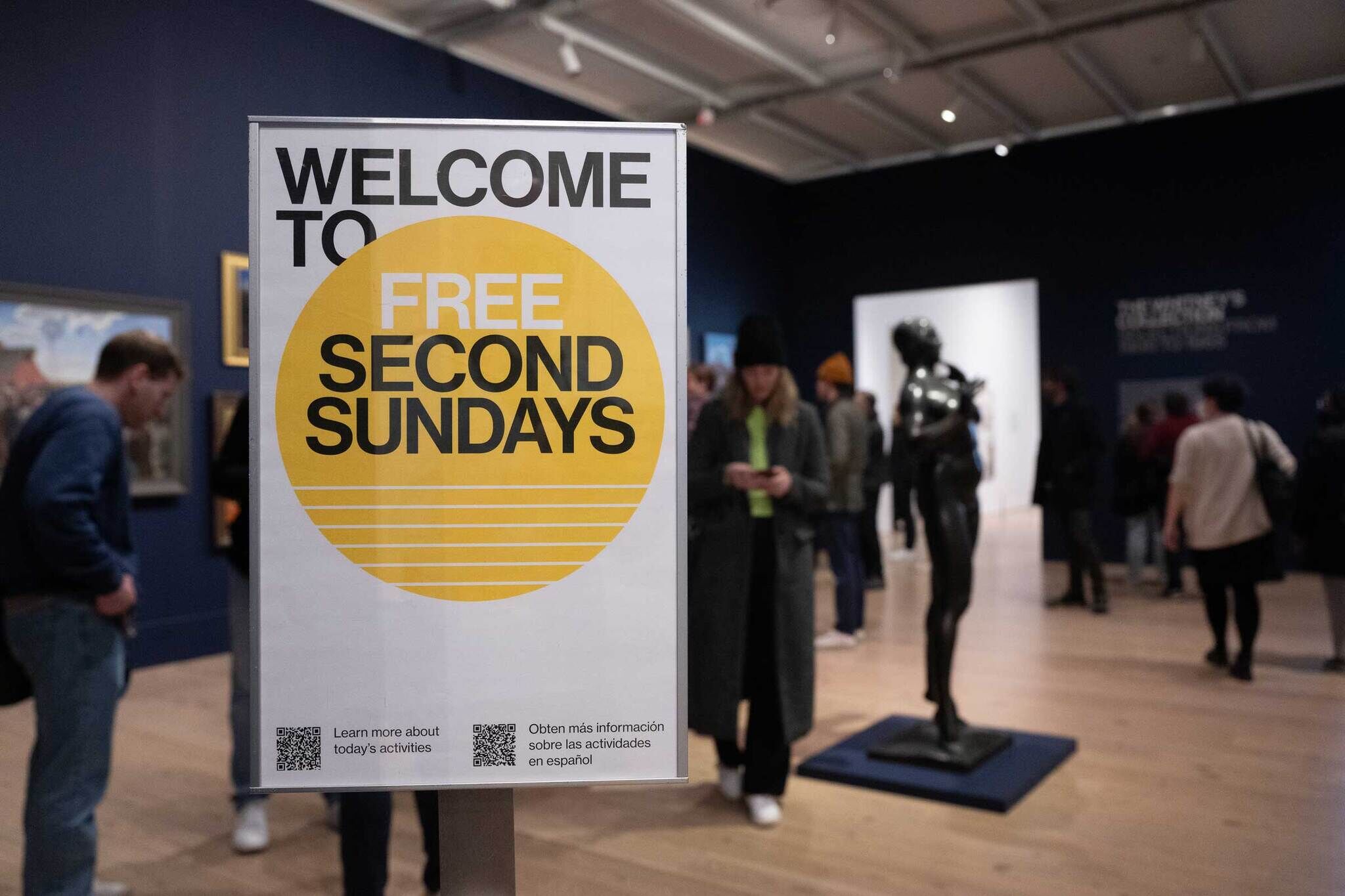 Sign reading "Welcome to Free Second Sundays" in a museum, with people viewing art and a statue in the background.