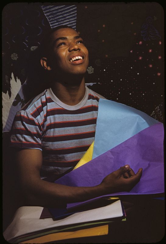 A person in a striped shirt smiles while holding colorful sheets of paper, looking upwards against a dark, patterned background.