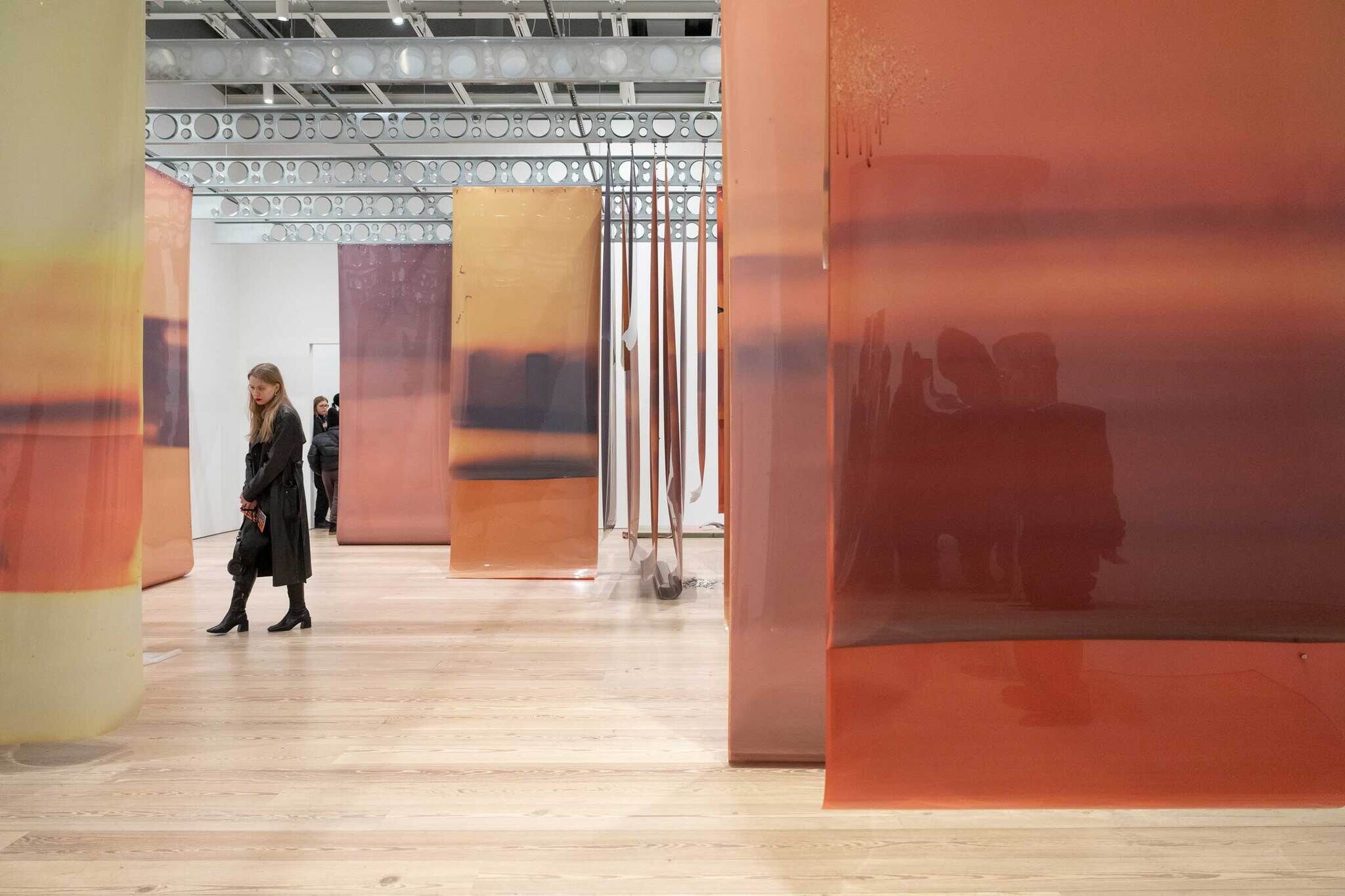 A woman in a black coat walks through an art installation with large, semi-transparent colored panels hanging from the ceiling.