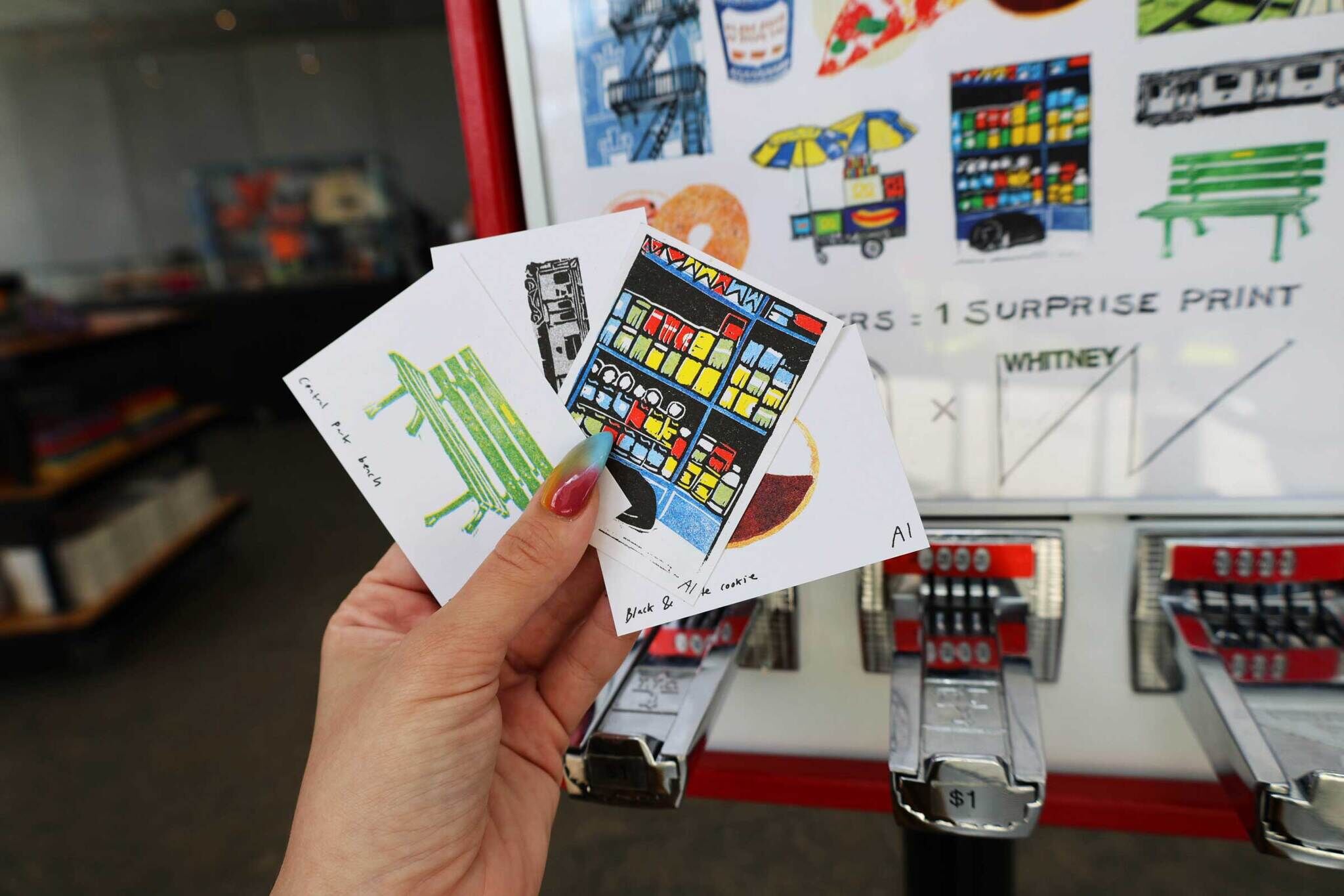 A hand holding three illustrated cards in front of a vending machine. The cards depict a green bench, a colorful store, and a cookie.