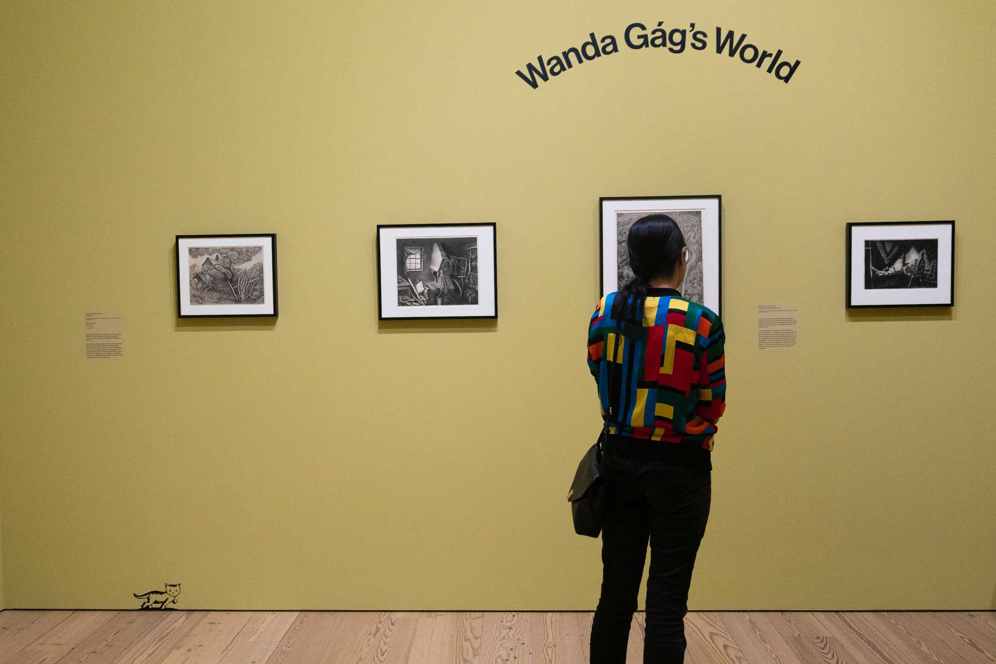 Person in a colorful shirt views framed black-and-white artwork in a gallery titled "Wanda Gág's World" with yellow walls and wooden floors.
