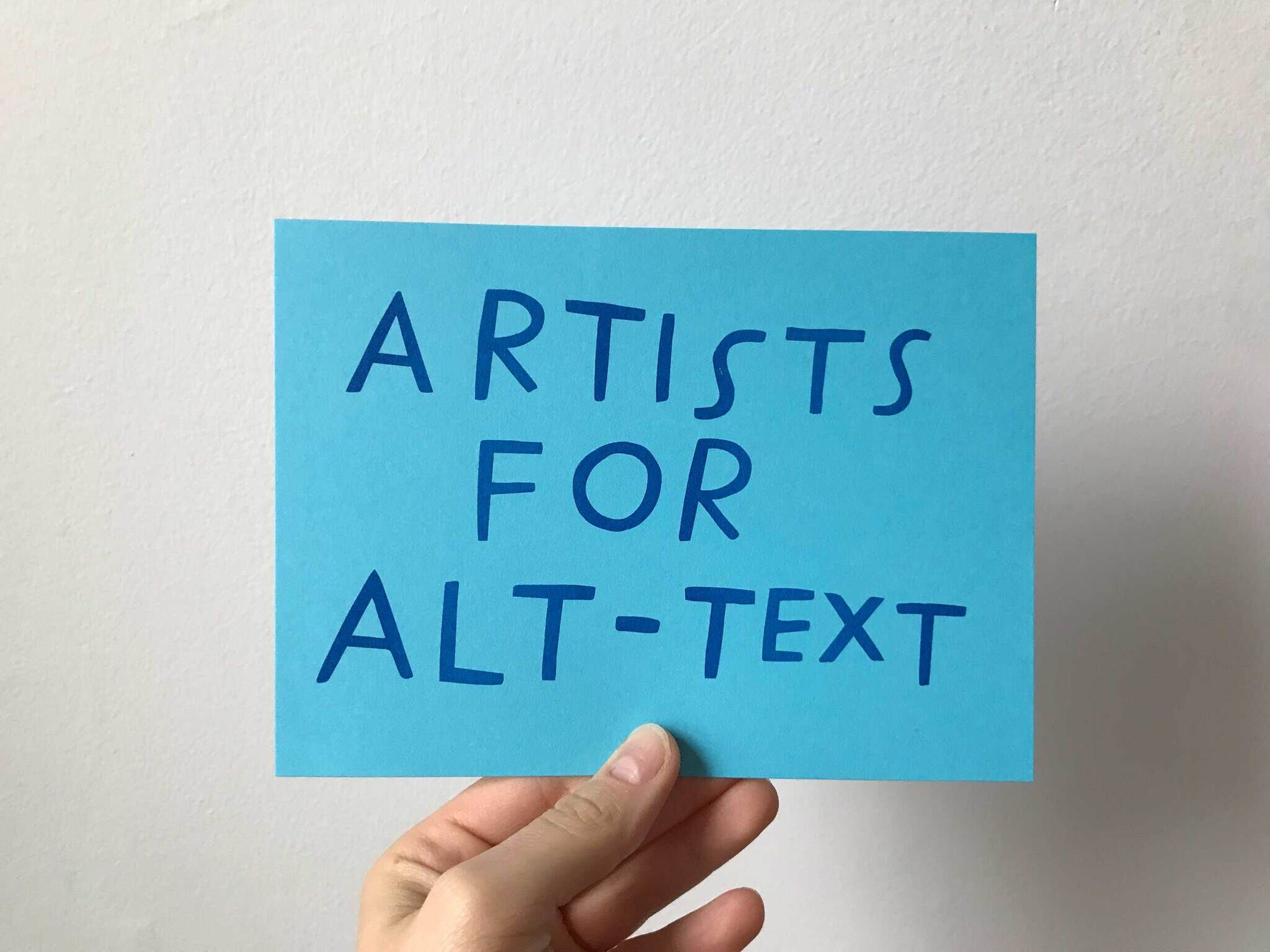 A turquoise postcard with contrasting navy, playful, handwritten capitals spelling, “Artists for Alt-Text.”. The words are boldly written and a slender white hand gently holds the postcard as if to say, “hey take a look!”.